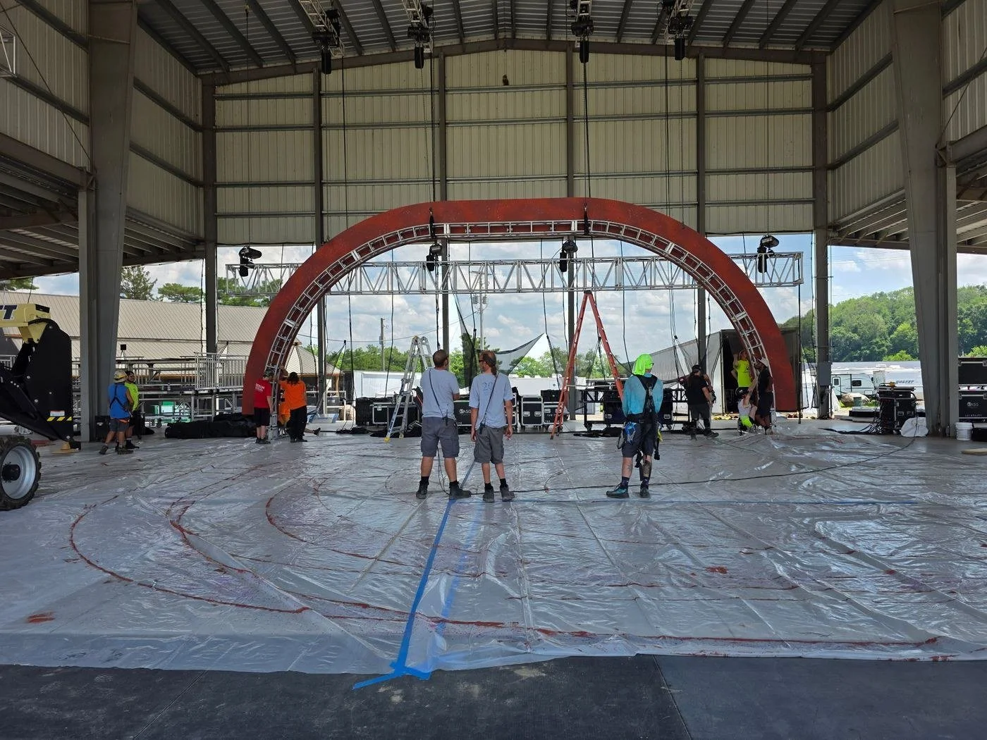 Crew laying out a stage floor under an open-air pavilion, with a large scenic arch flown on chain hoists