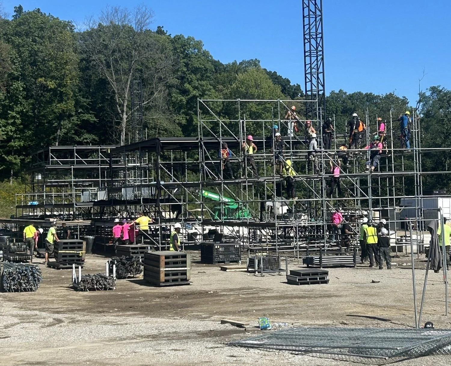 Crew in hard hats and harnesses building a scaffold festival stage, with deck and pipe staged on the ground