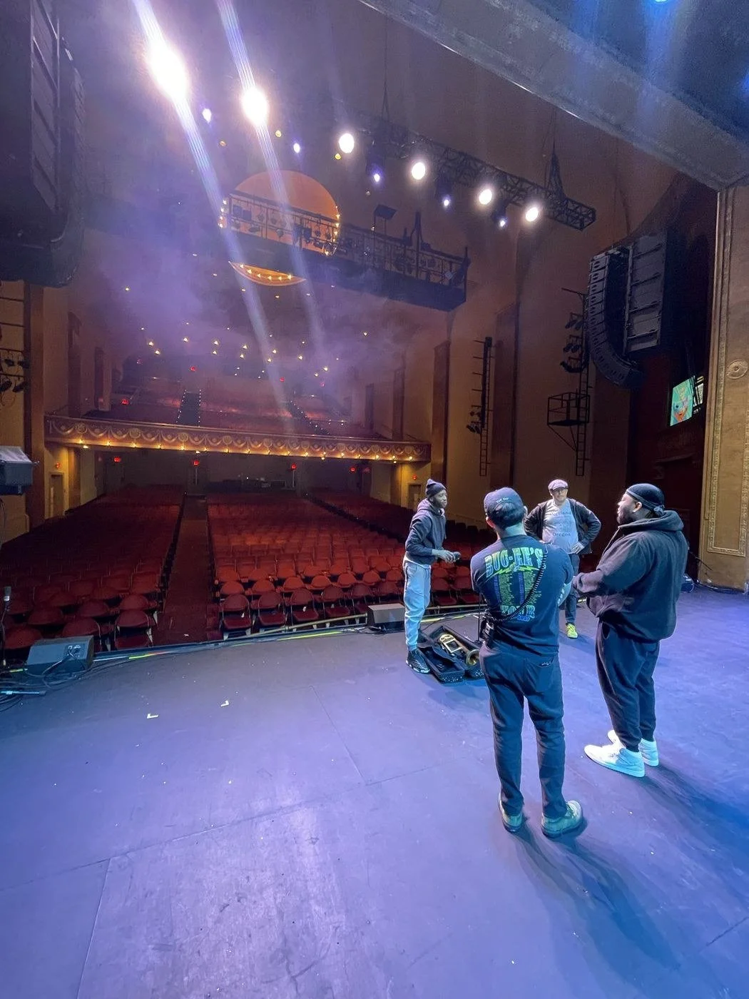 Crew and band on a theater stage, with line arrays and rows of red seats behind them