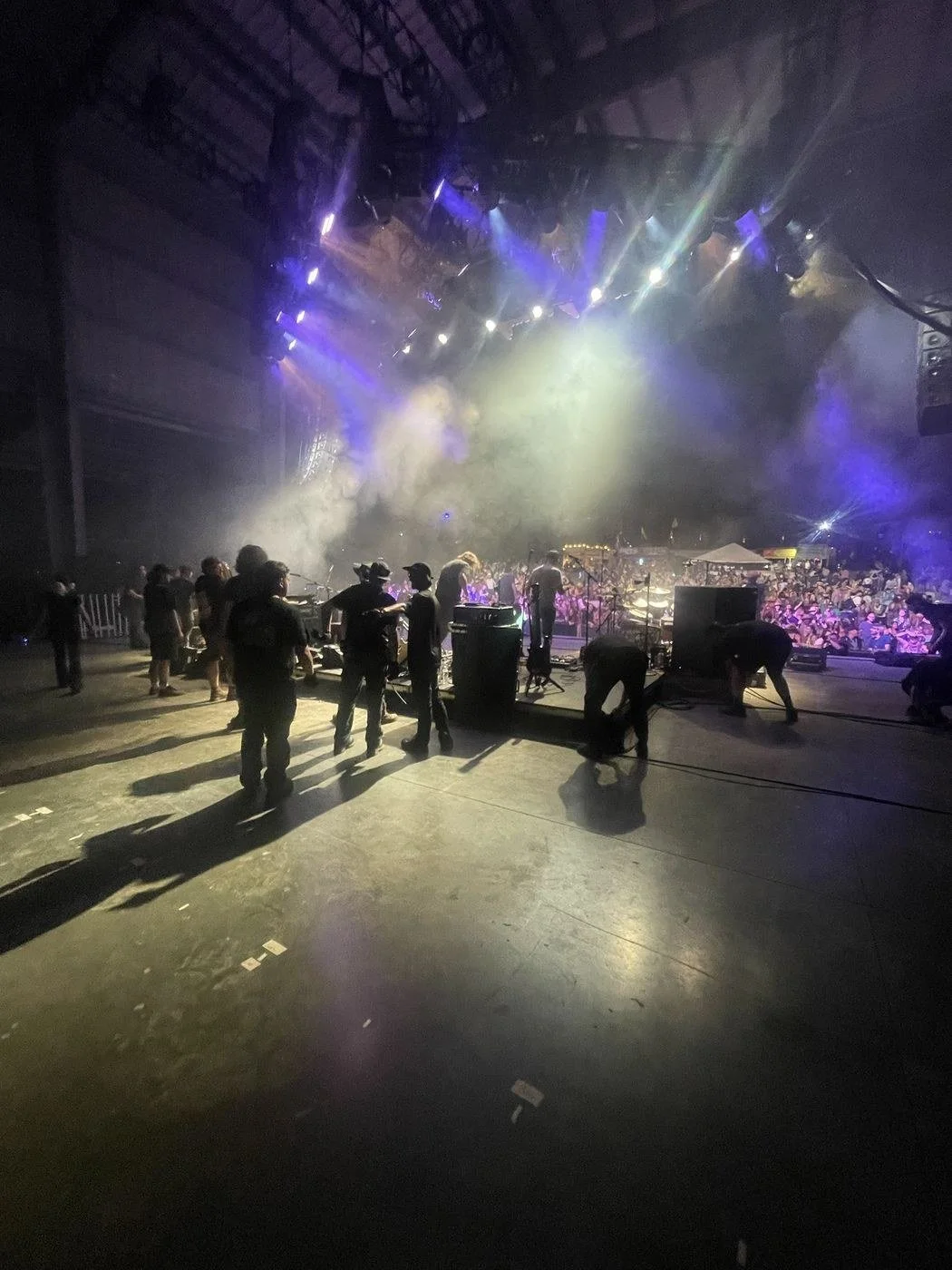 Stagehands working on a festival stage under moving lights, with the crowd beyond