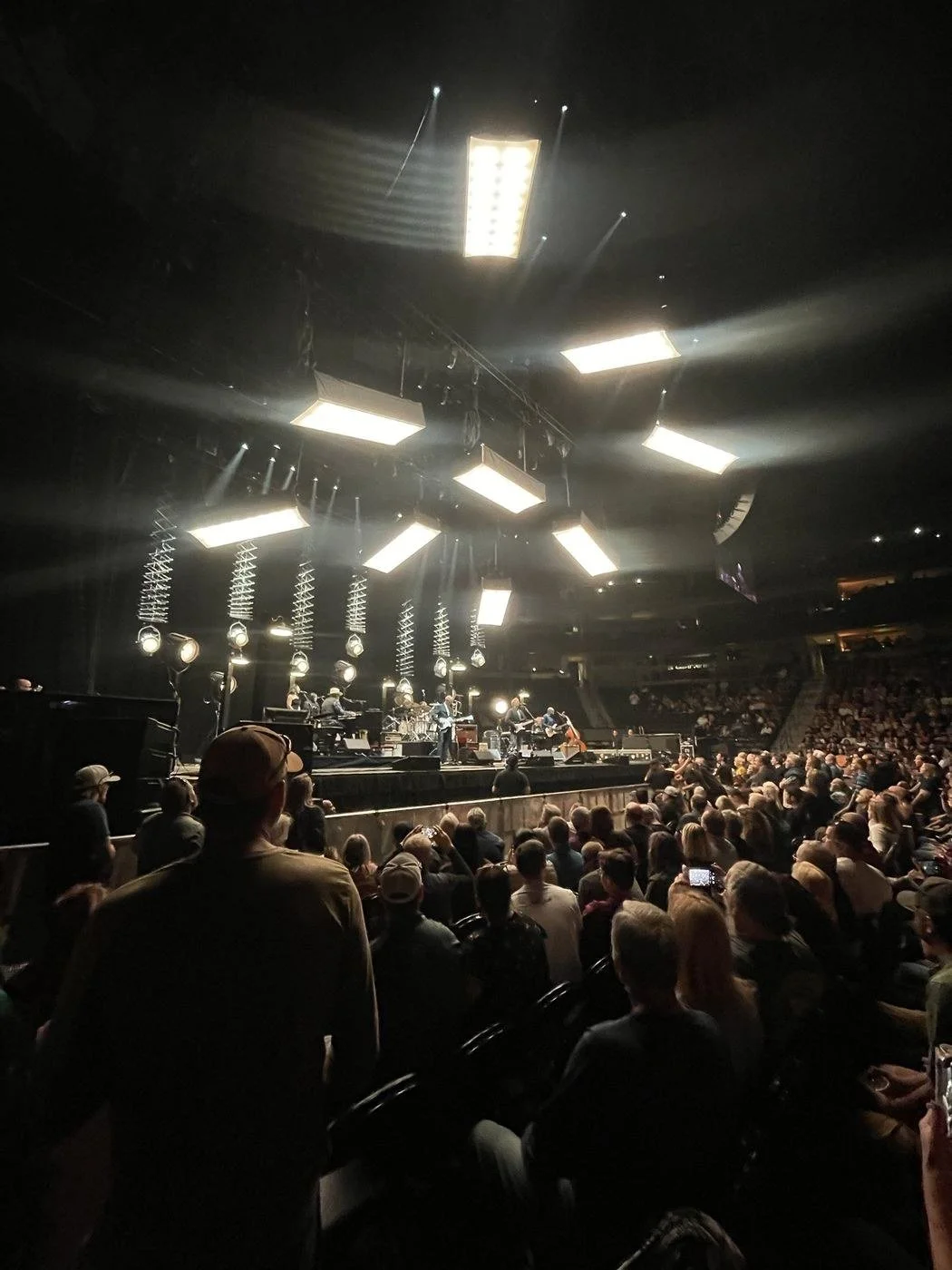Arena concert with overhead lighting pods and a full crowd on the floor