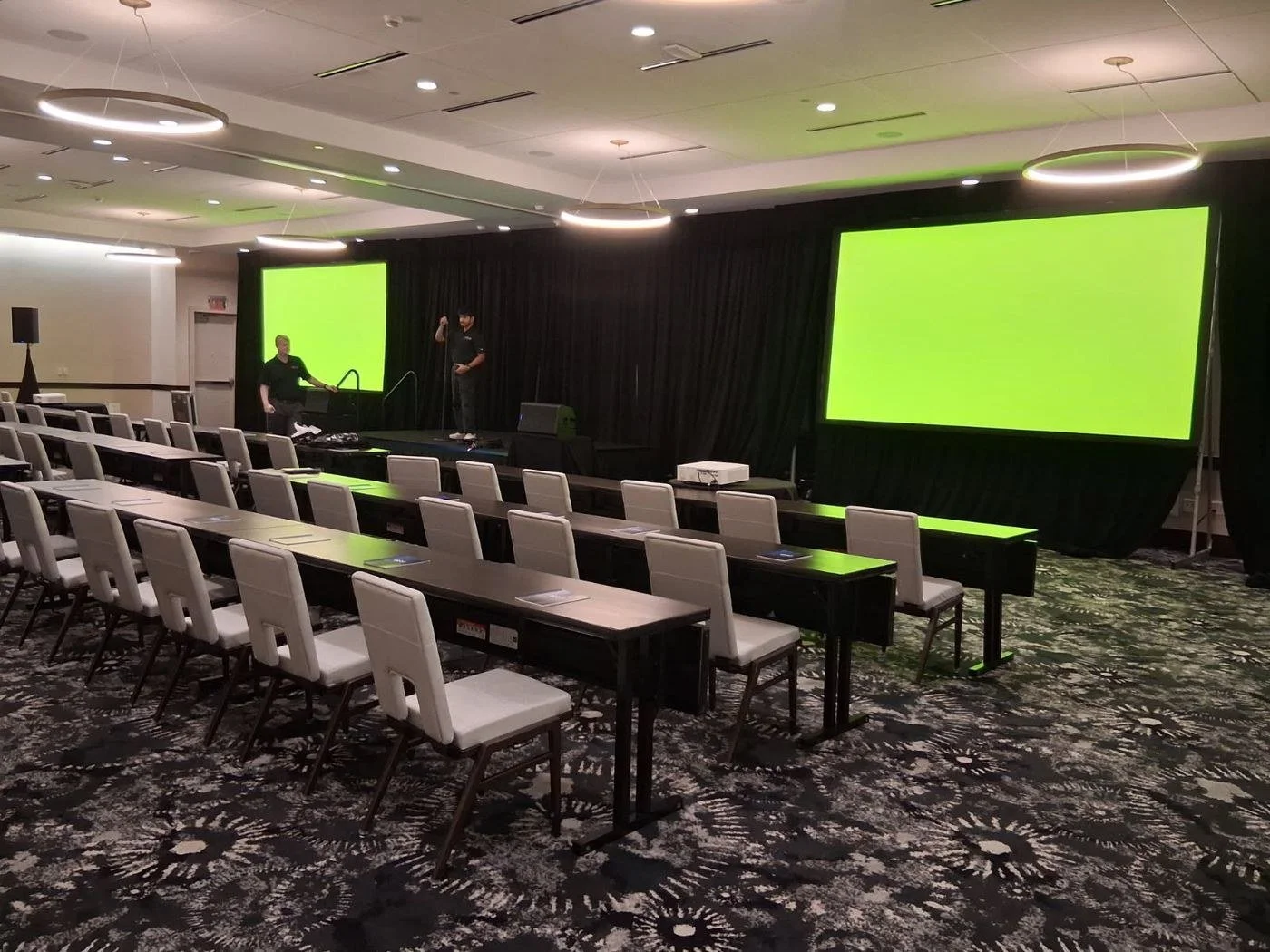 Hotel ballroom set for a corporate session with two projection screens, a stage and black drape