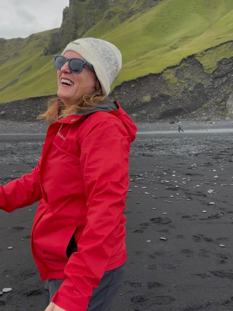 Samantha laughing in a red rain jacket and knit hat on a black sand beach in Iceland