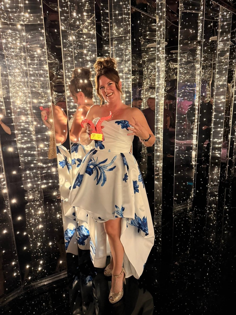 Samantha Robichaud in a white and blue floral gown, holding a Virgin Voyages award in a glittering mirrored room