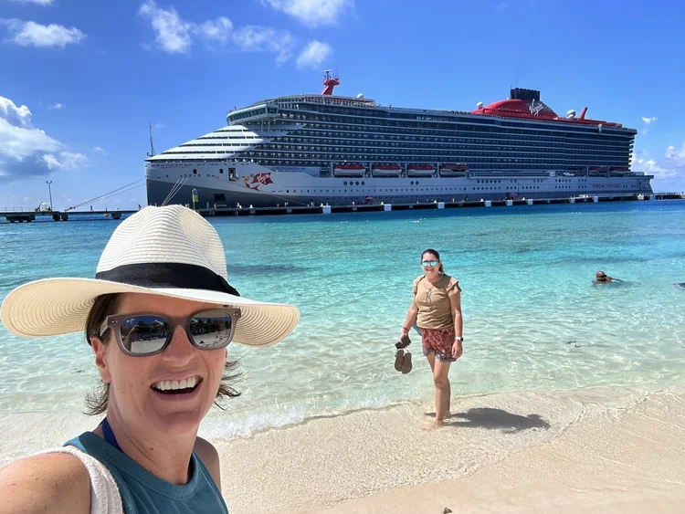 Samantha in a white sun hat smiling on a beach, with the Virgin Voyages ship Scarlet Lady docked behind her