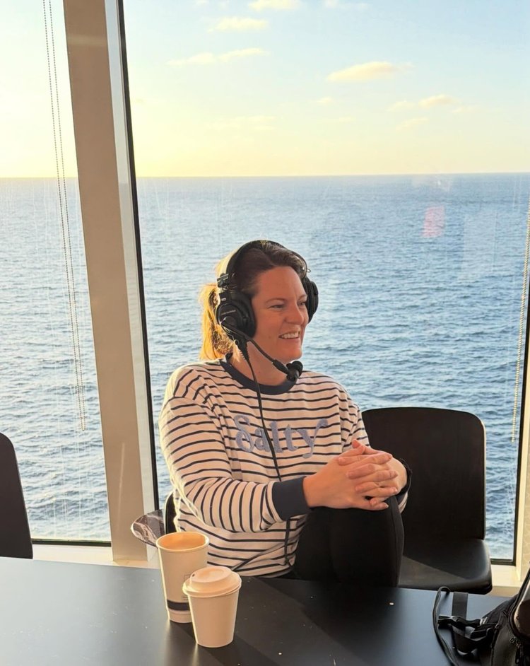 Samantha Robichaud laughing in a headset and a striped Salty sweatshirt, recording at a table by a window over the open sea