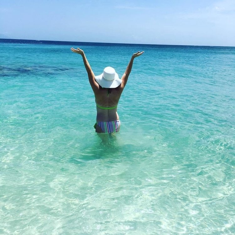 Samantha in a white sun hat, arms raised, wading into clear turquoise water