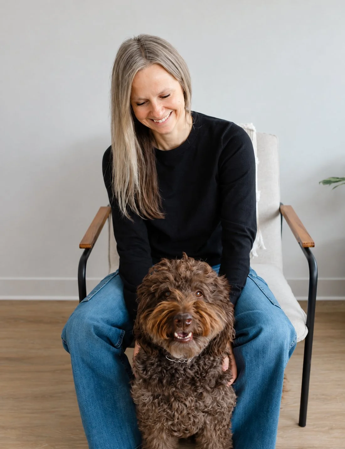 A smiling woman seated in an accent chair with her curly brown doodle