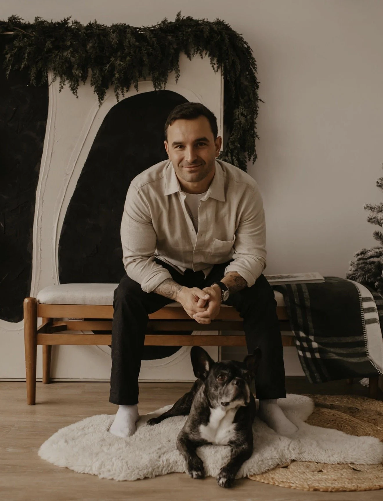 A man seated on a wooden bench with his French bulldog, styled with garland and a flocked tree for a holiday session