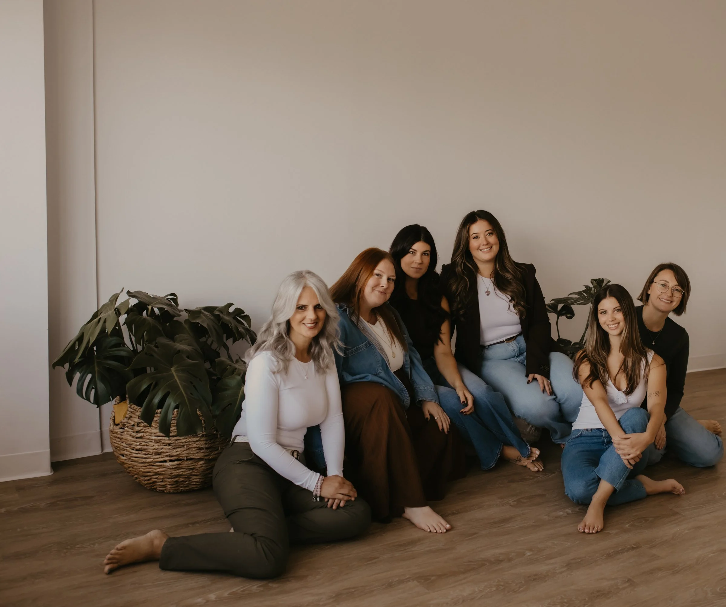 A group of six women sitting together on the studio floor beside a large monstera plant