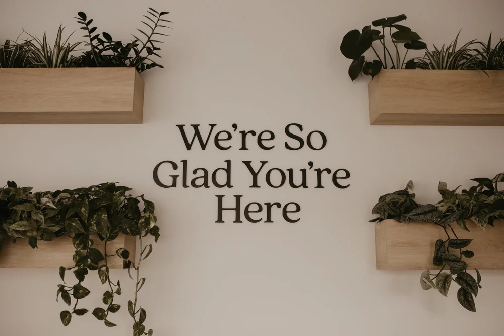 A wall sign reading We're So Glad You're Here framed by wooden planters