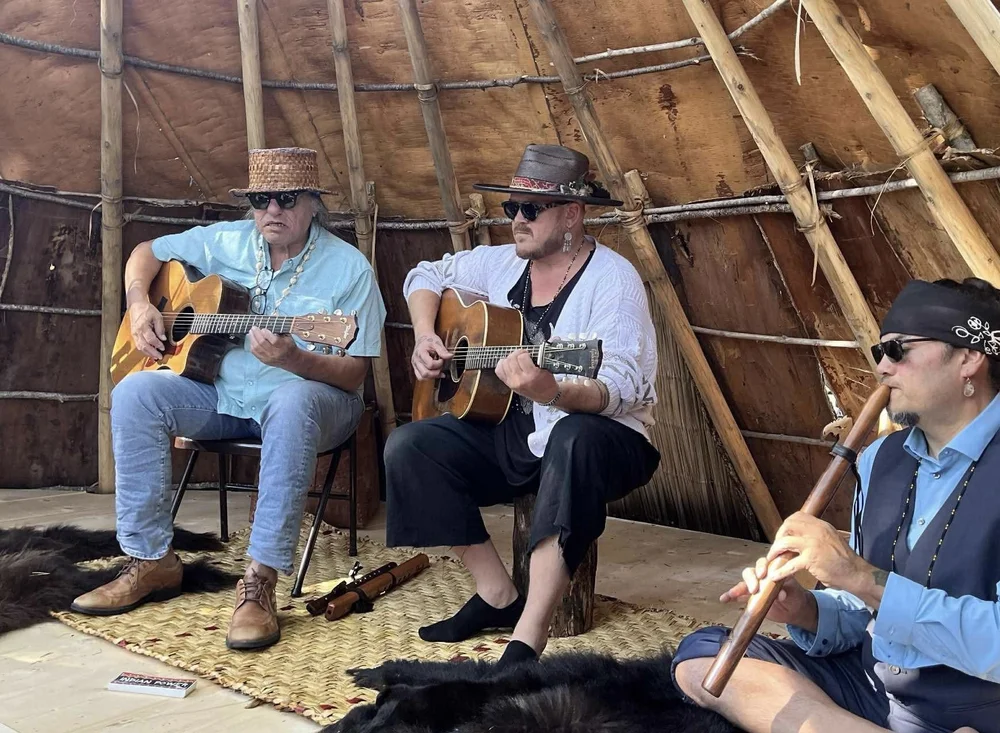 Three musicians performing together inside Birch House on Madeline Island, surrounded by the natural timber framework of the traditional bark house.