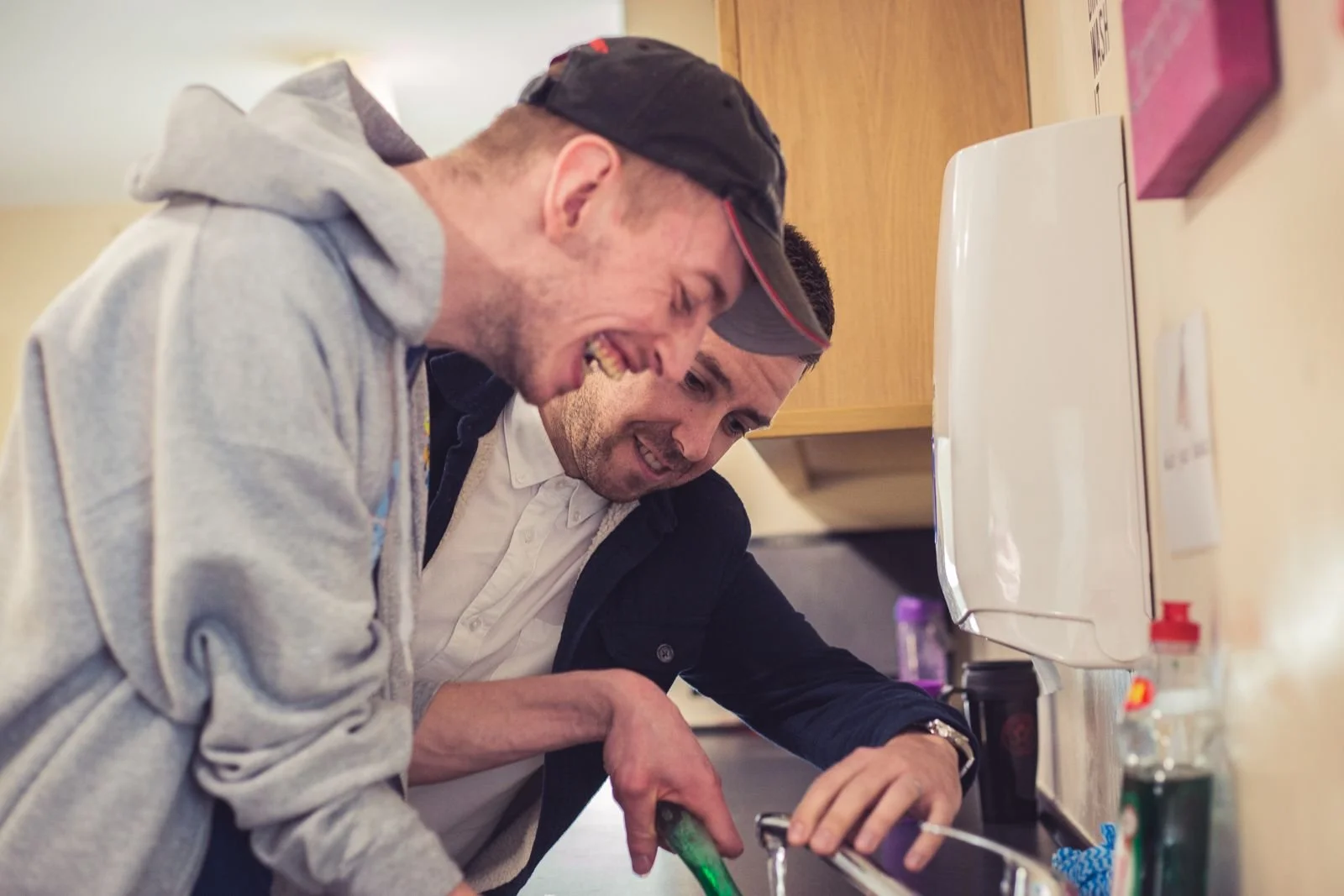 A support worker and a man in a cap laughing together while washing up