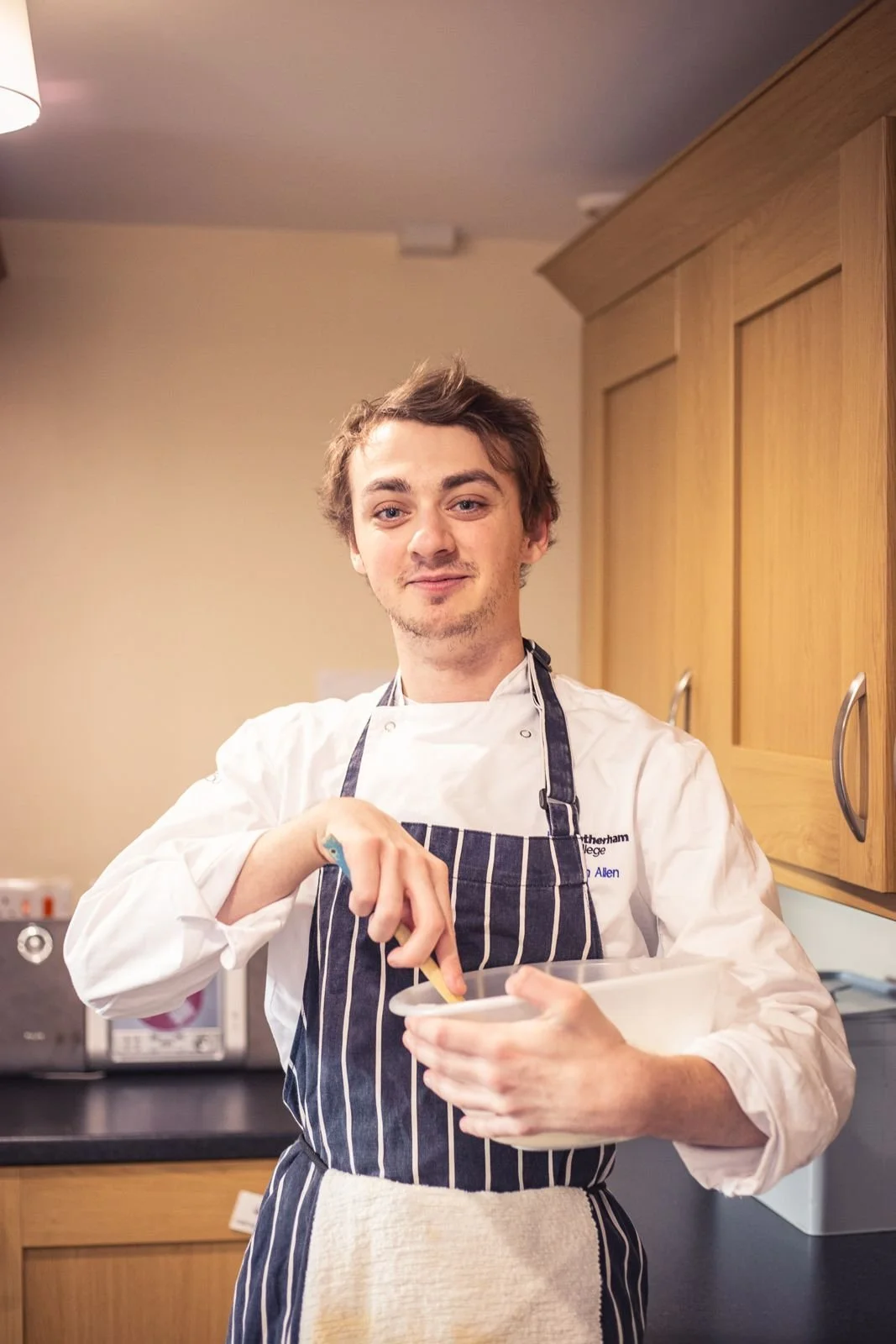 A young man in chef whites stirring a mixing bowl in a kitchen
