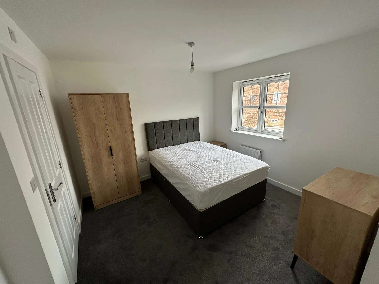 A furnished double bedroom with a grey bed, wooden wardrobe and window