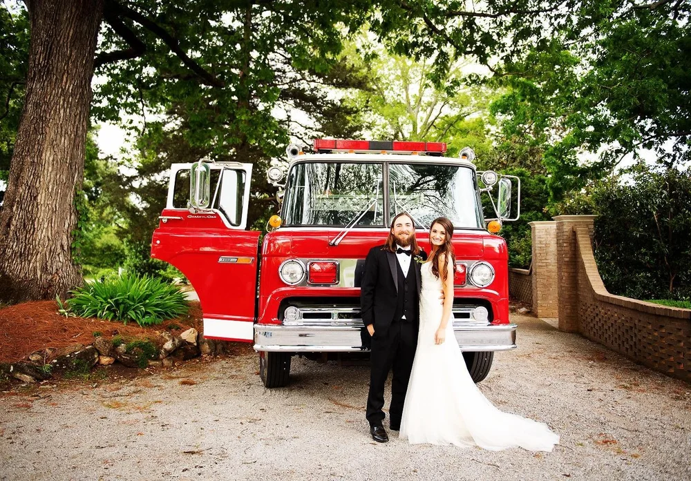 A wedding couple standing in front of the red-and-white Engine 706 firetruck under trees.