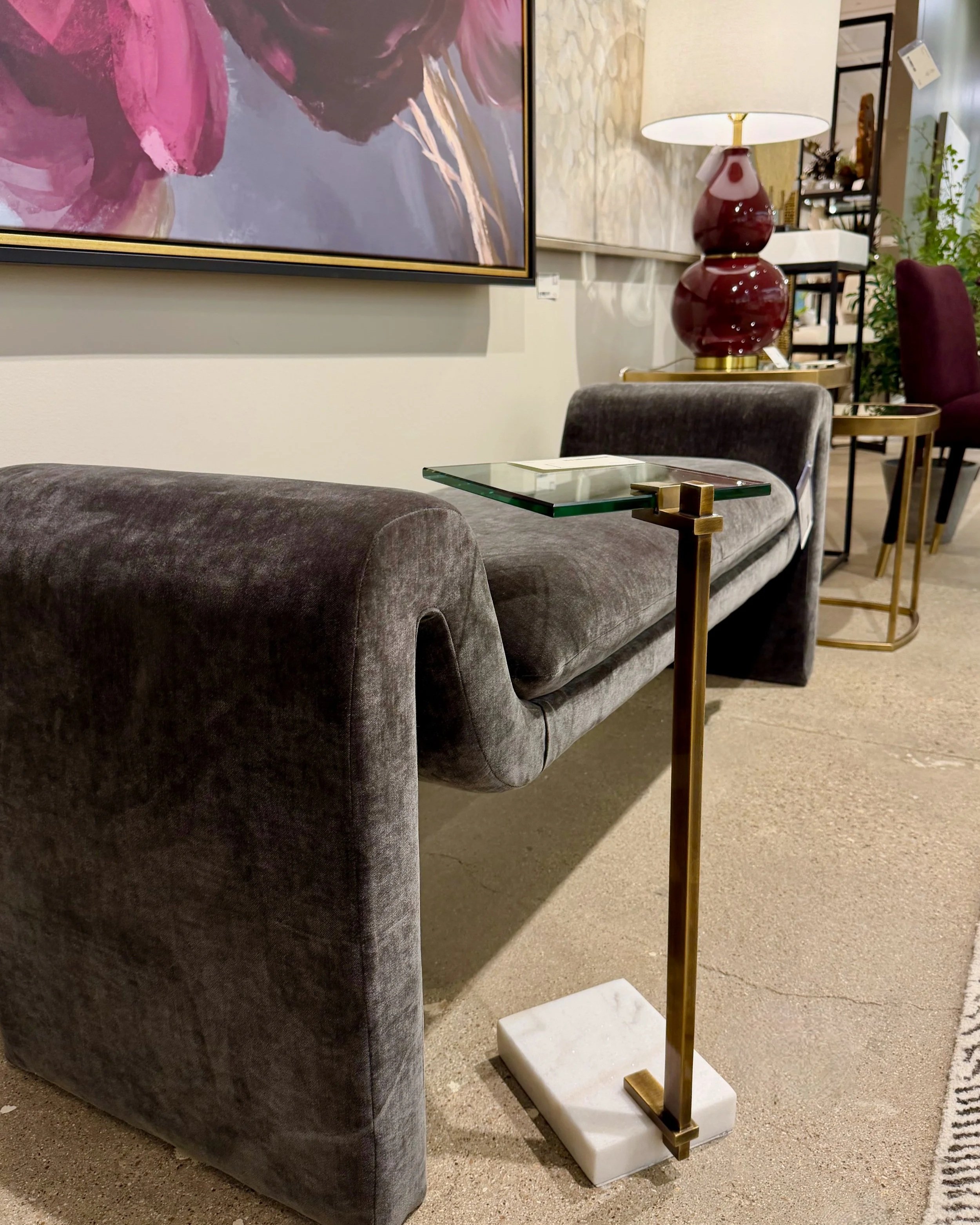 This image shows a grey upholstered bench in a modern style paired with a glass topped drink table. Plum colored art and a lamp are in the back ground.