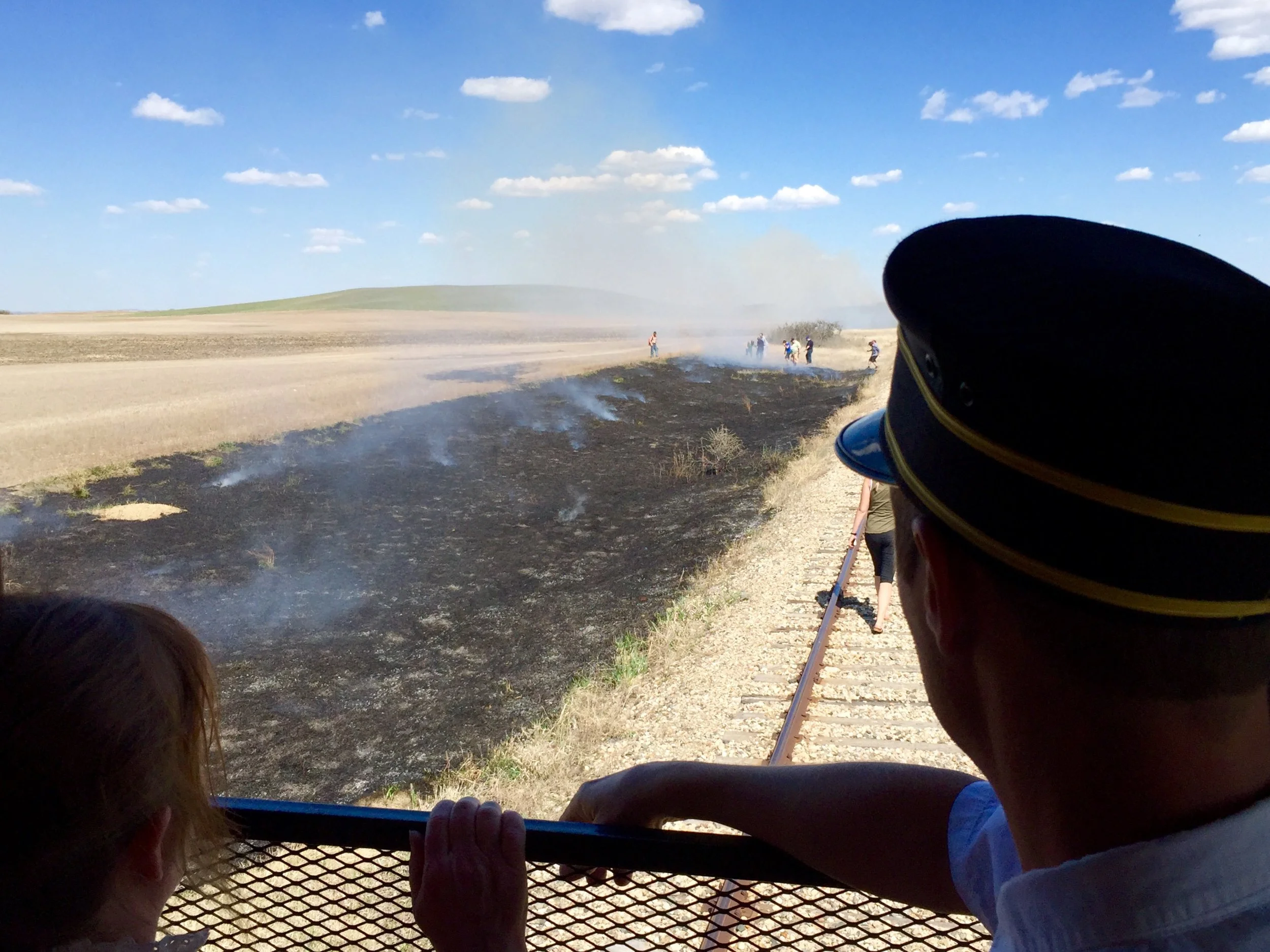 A view from the back of the Southern Prairie Railway train. A little girl (left) and the train conductor (right) frame the bottom of the photo along with a metal railing. Beyond them is a long patch of burning ditch grass, which is black and smoking.