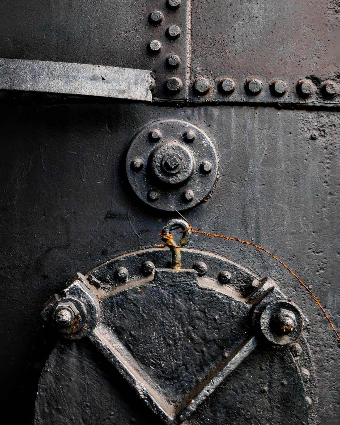 &ldquo;Ironside&rdquo; &copy; 2025 Skip Hunt

Riveted metal panels are arranged with precise geometric patterns, featuring large bolts and a dark surface that suggest industrial machinery. A thick wire, coiled and slightly rusted, hangs from one of t