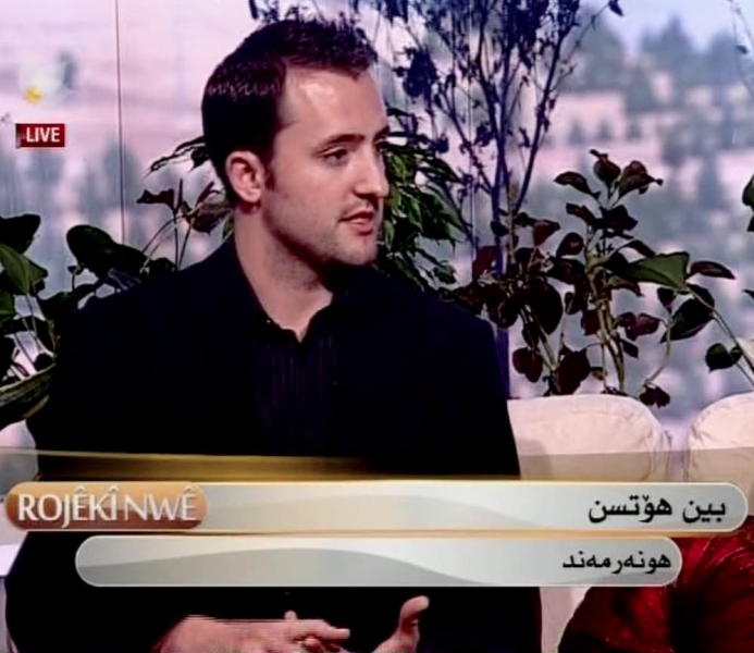 A man with short dark hair and a black shirt sitting on a television show set, with a background of plants and a cityscape. There is a live broadcast indicator and text in a non-English language.
