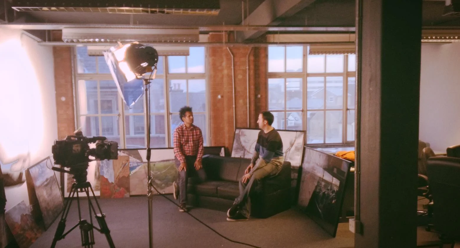 Two people having a conversation in a studio apartment, with large windows, paintings leaning against the wall, a professional light and camera on tripods.