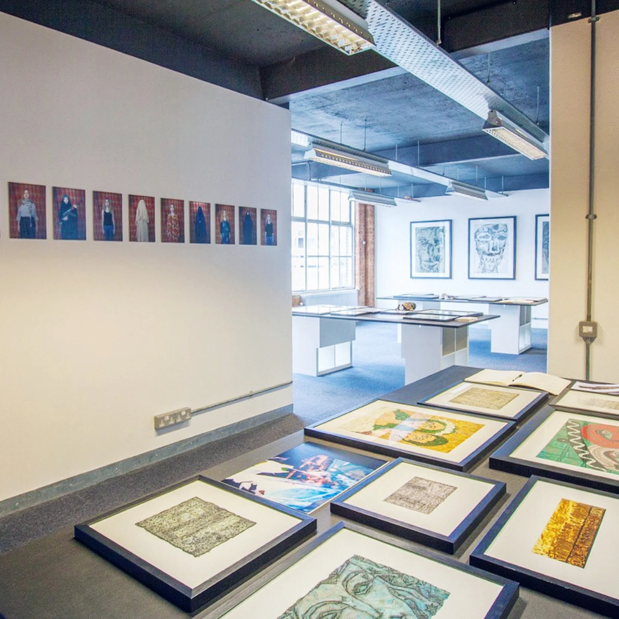An art gallery with framed artwork displayed on walls and tables, natural light coming through large windows.