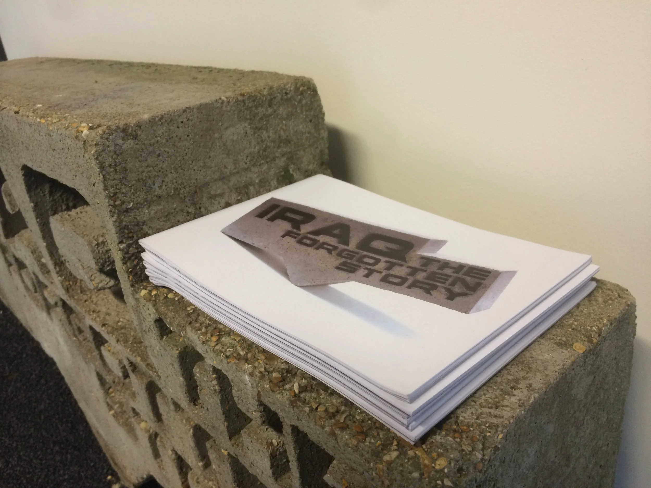 Stacks of paper with a faded black and white image that reads "IRAQ THE FORGOTTEN STORY" placed on a rough concrete block against a plain wall.