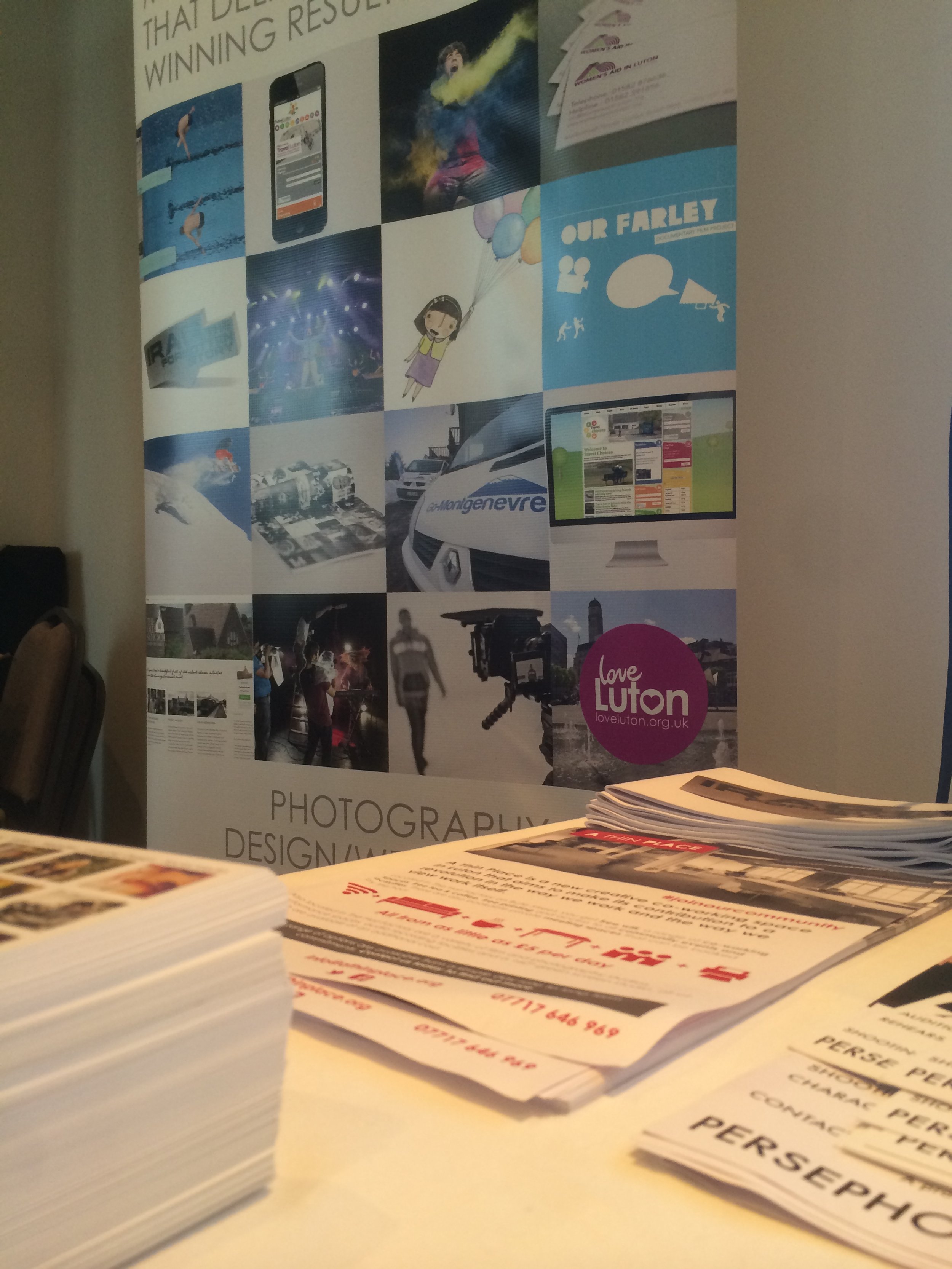 A table with stacks of papers and flyers in front of a large poster featuring various images related to photography, design, and media. The poster includes pictures of people, cars, a computer screen, and graphics, with some logos and text.