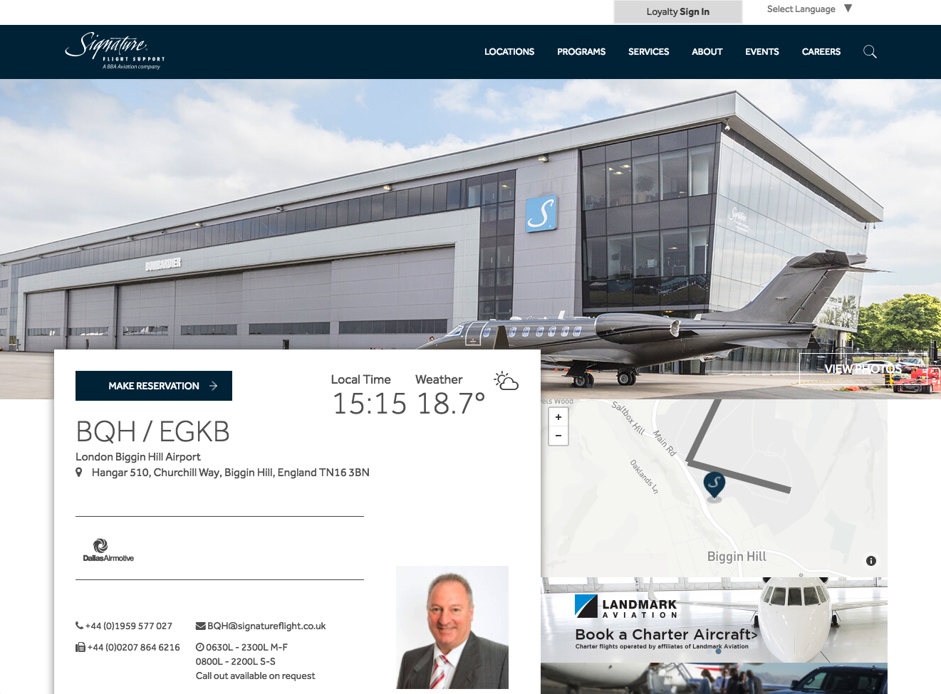 Exterior of a modern private jet hangar at London Biggin Hill Airport with a sleek private jet parked outside, featuring a large blue signage with a white 'S' logo, and additional airport information visible on the screen.
