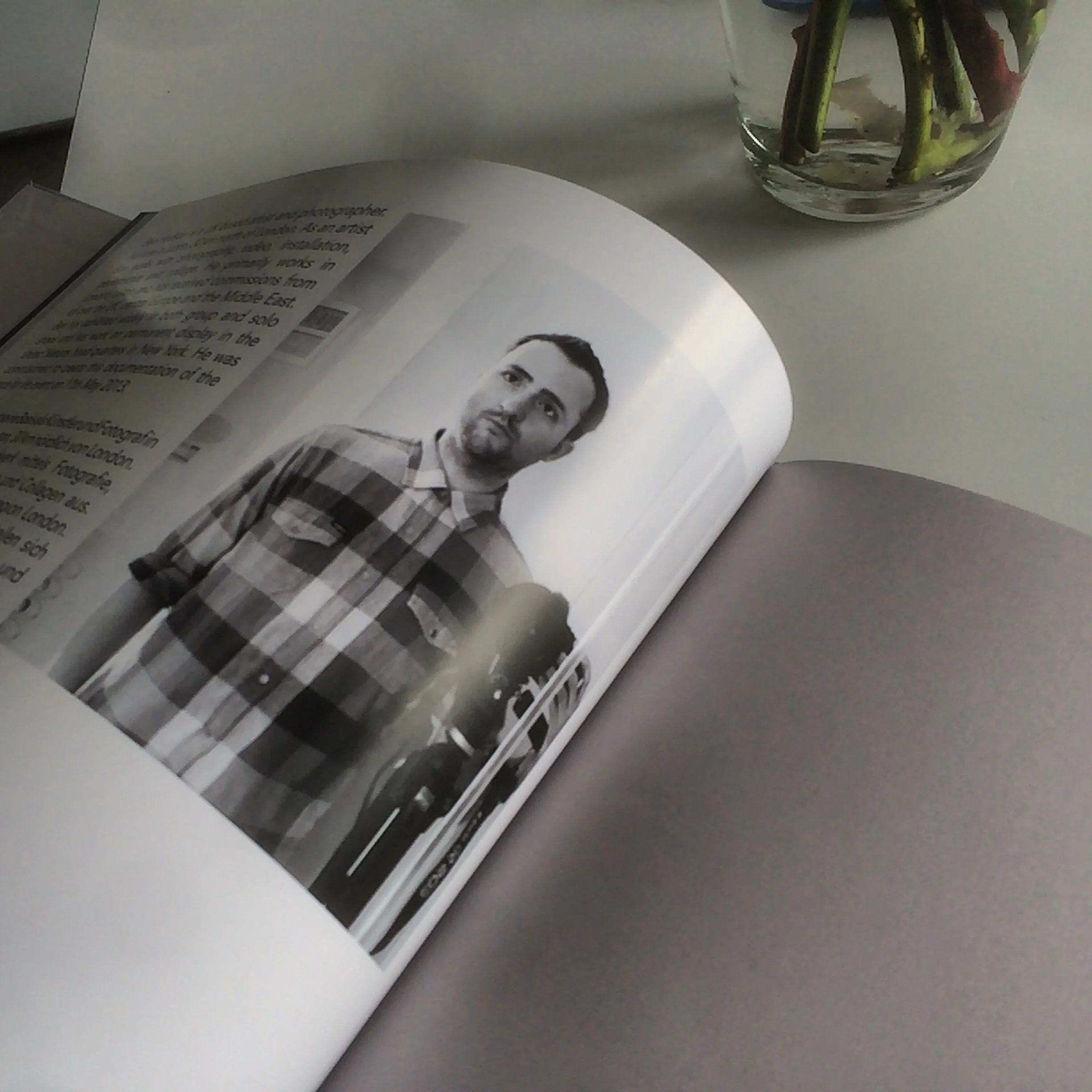 Open magazine with a black-and-white portrait of a man with a short beard, wearing a plaid shirt, on a table next to a glass vase with flower stems.
