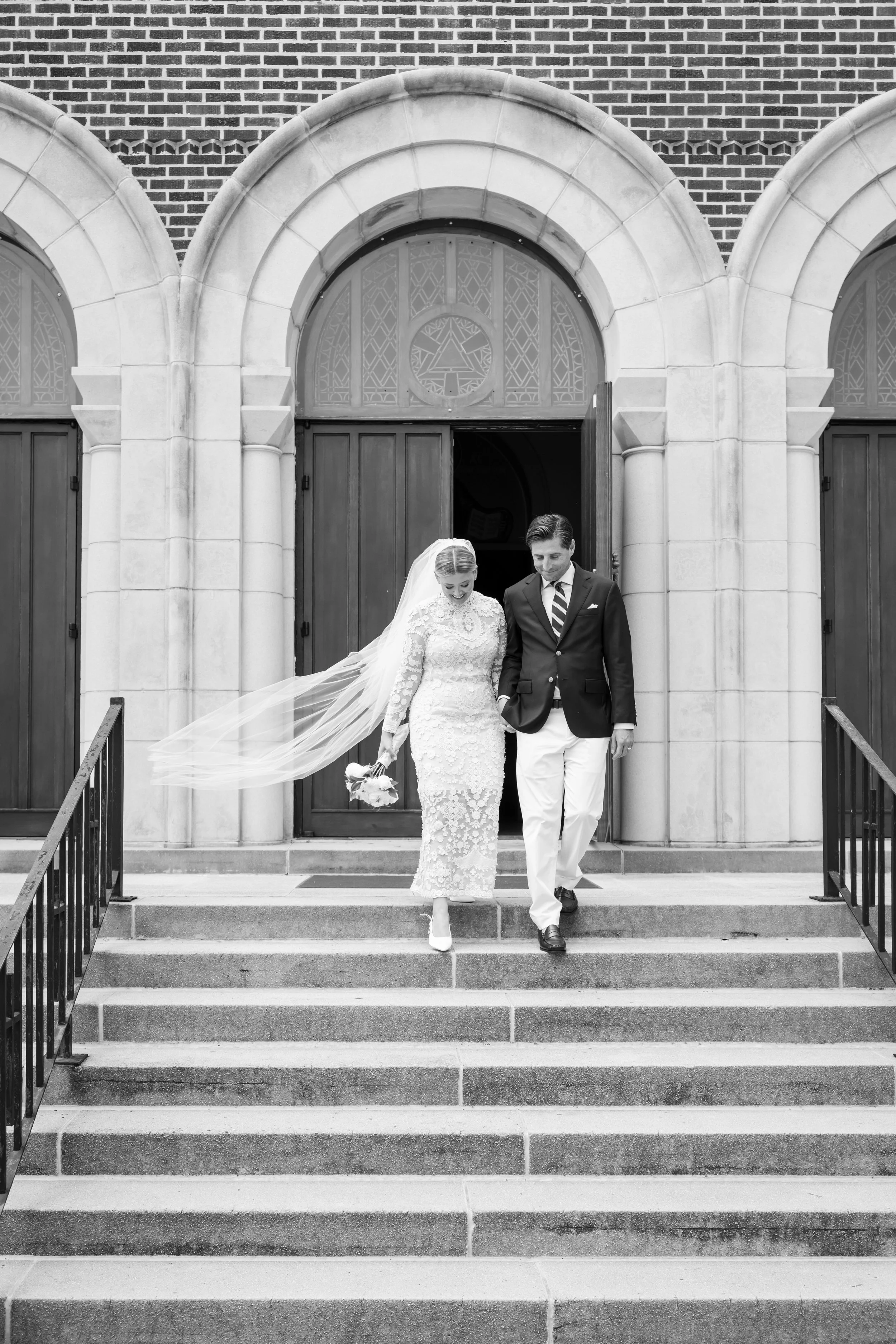 black and white of Just married couple exiting Charleston Greek church while brides long veil flows