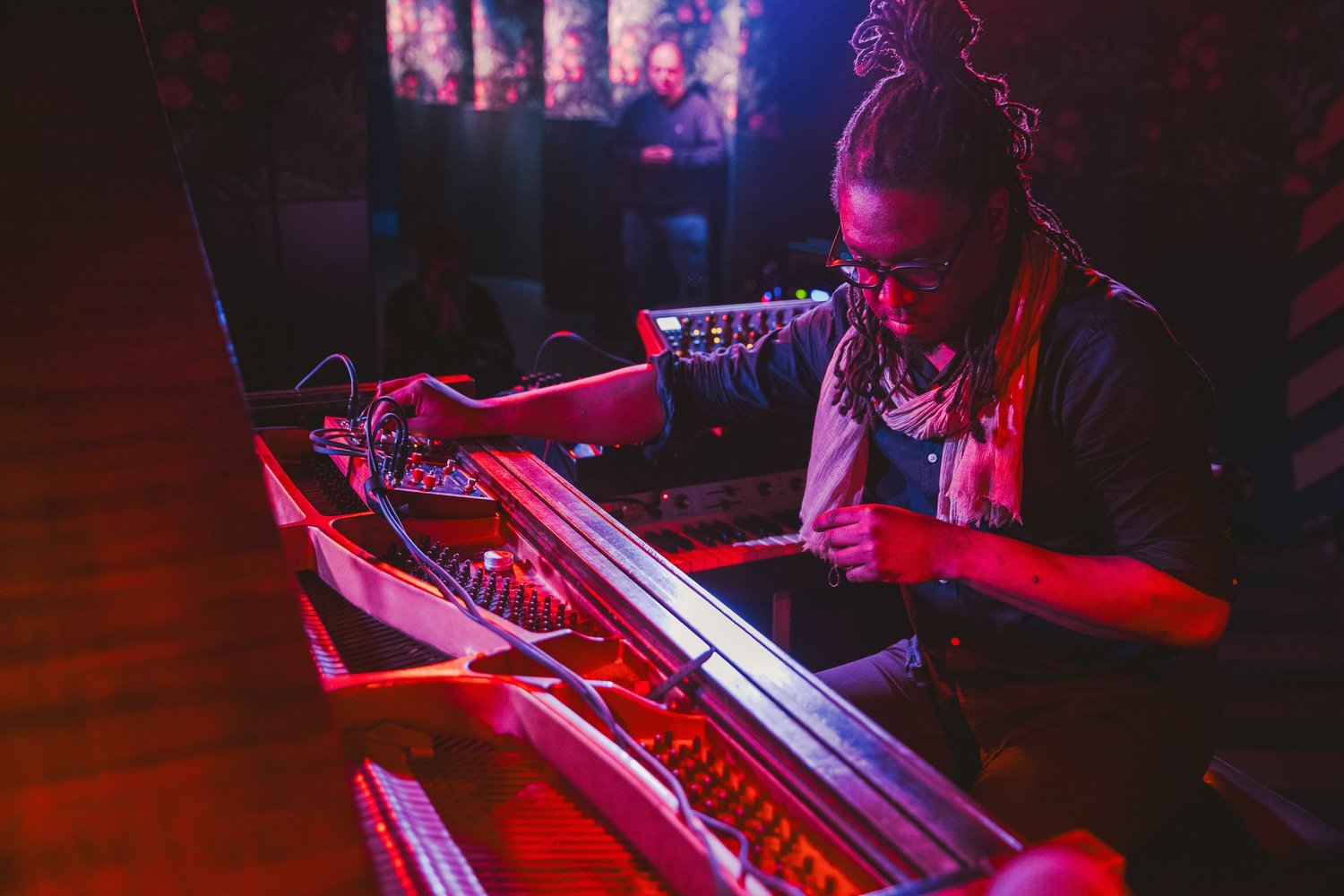 George Burton at an open piano wired with electronics, in red stage light, Bemis Center