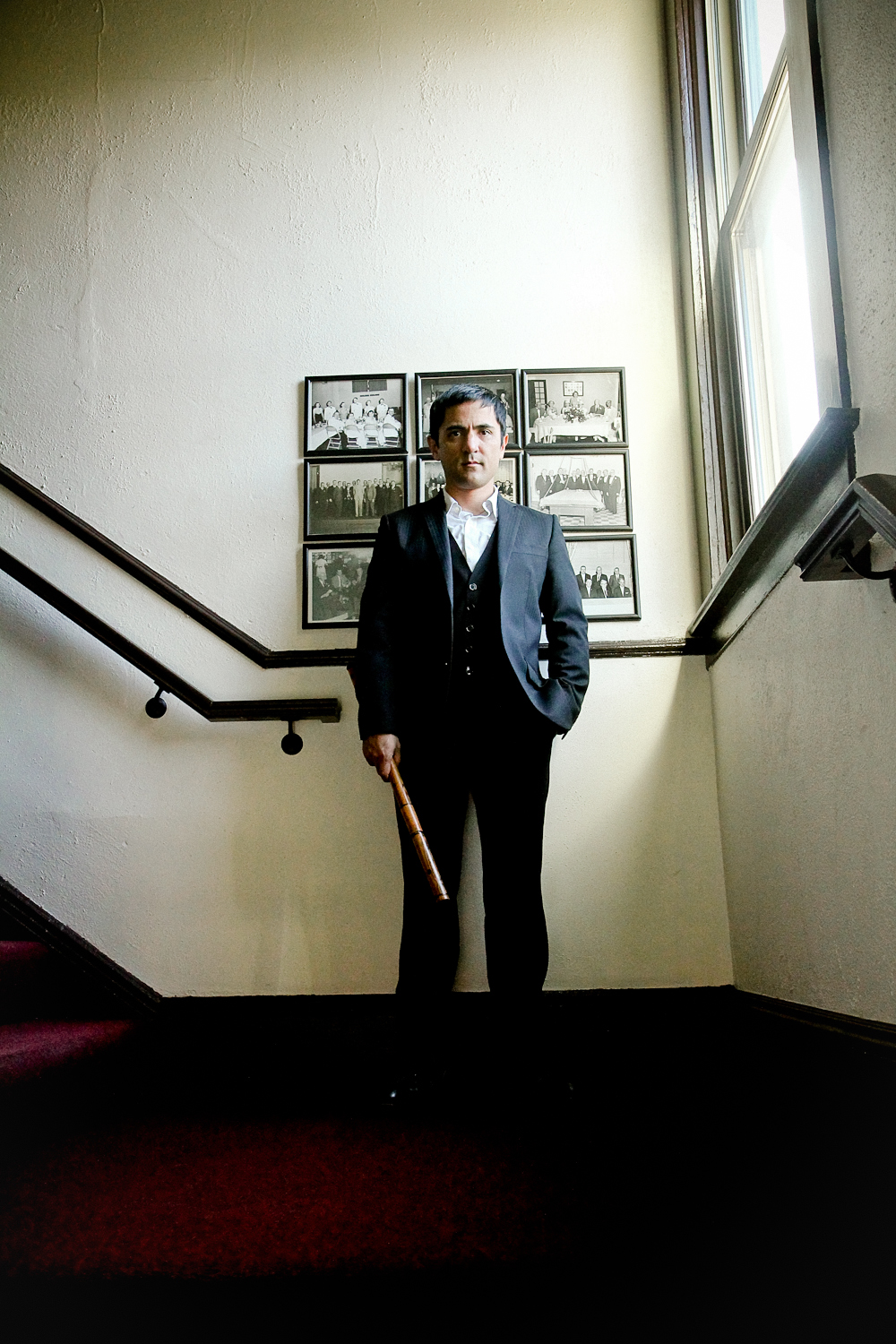 A man in a black suit standing on a staircase landing holding a wooden instrument, with black and white group photos on the wall behind him and a large window to his right.