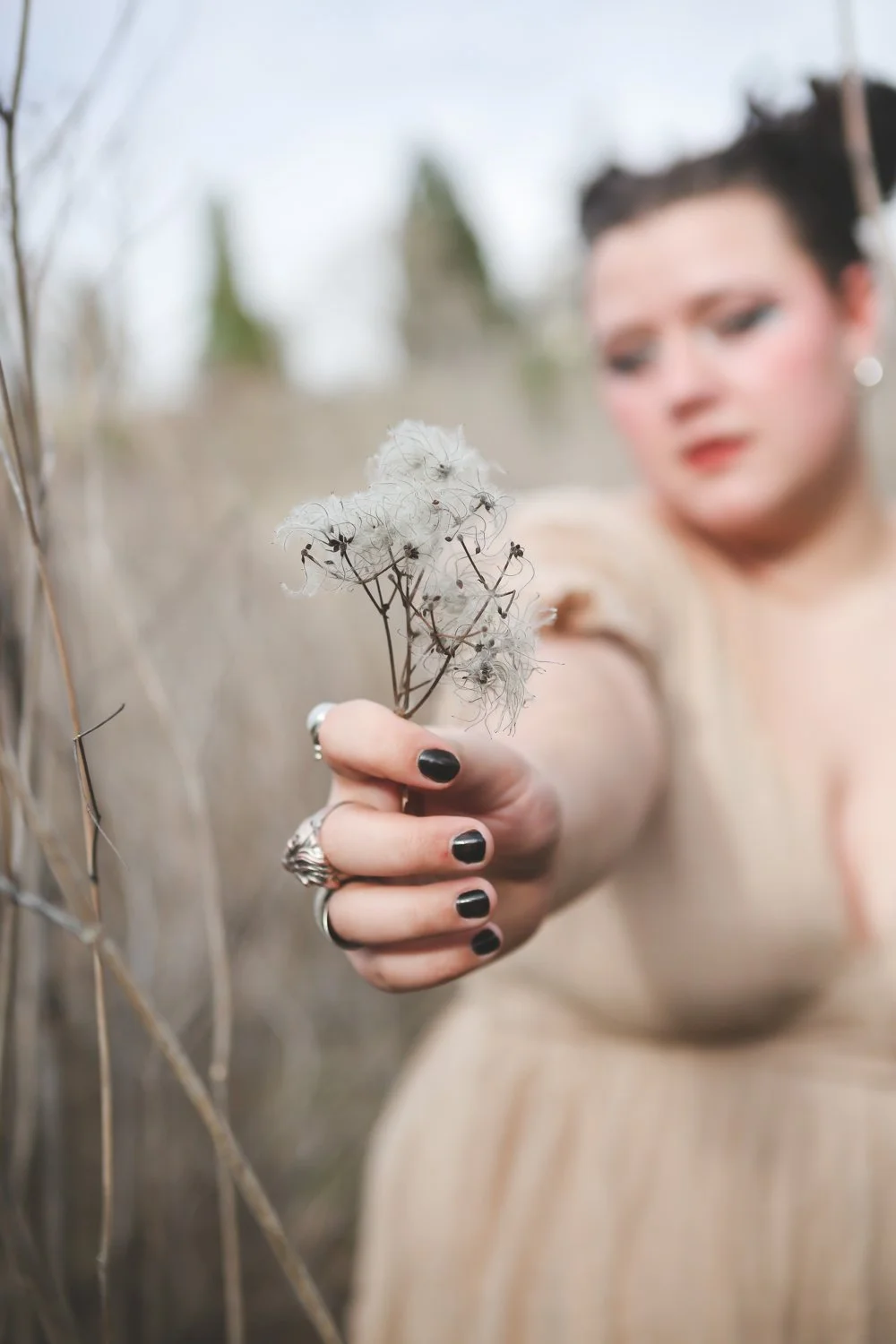 Woman with short dark hair and makeup, wearing a beige dress, holding a dandelion seed head with black nail polish and rings, standing outdoors in a field.