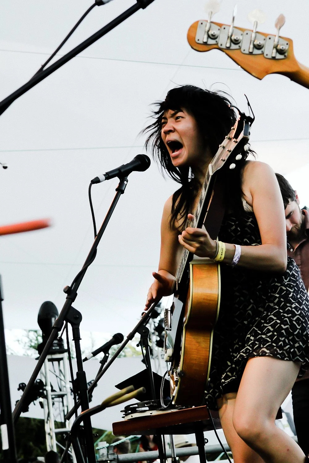 Female musician singing and playing guitar at a live outdoor concert with microphone and musical equipment.