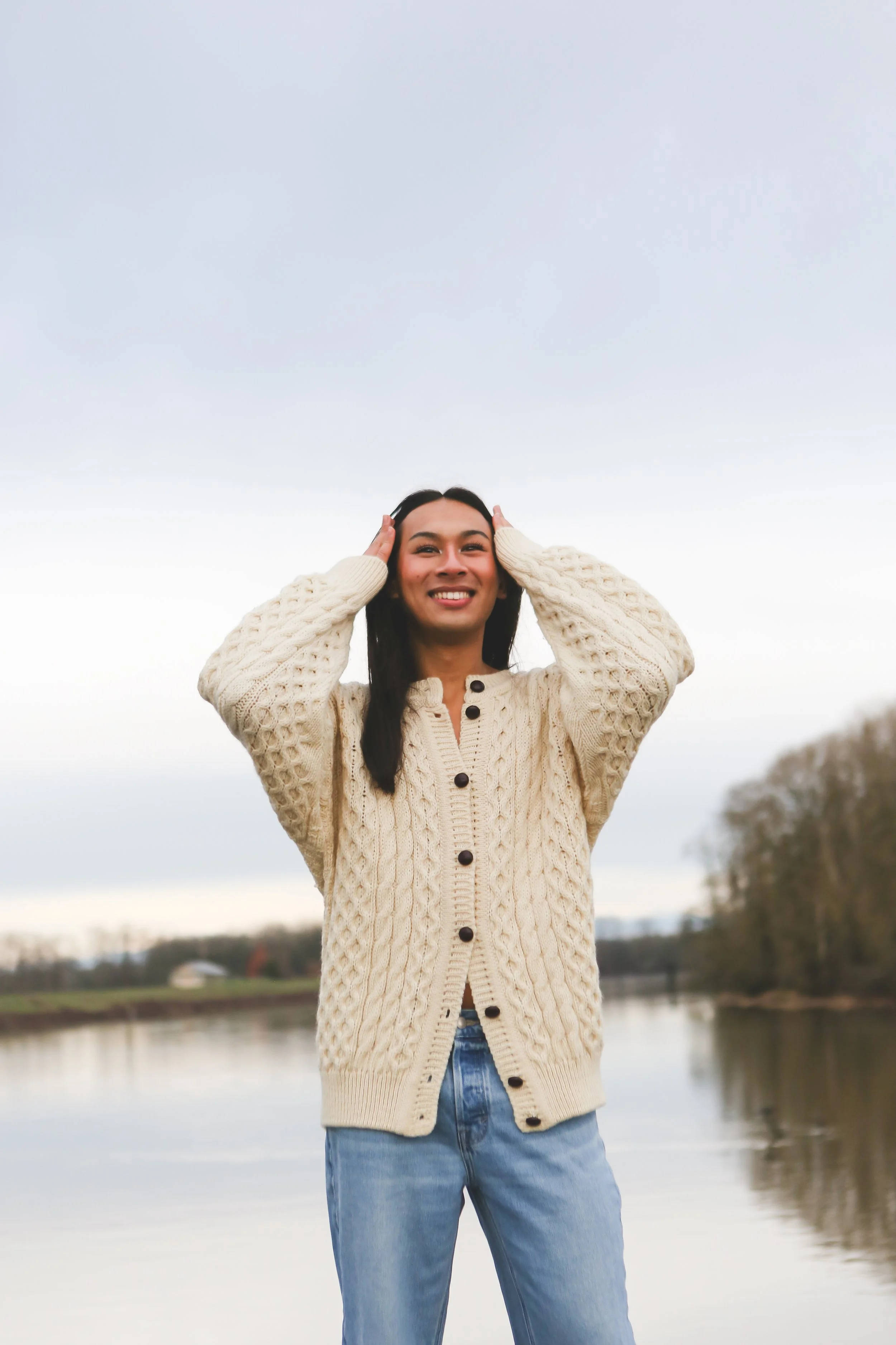 A woman wearing a beige cable-knit sweater and jeans standing outdoors near a body of water, smiling and holding her hands on her head.