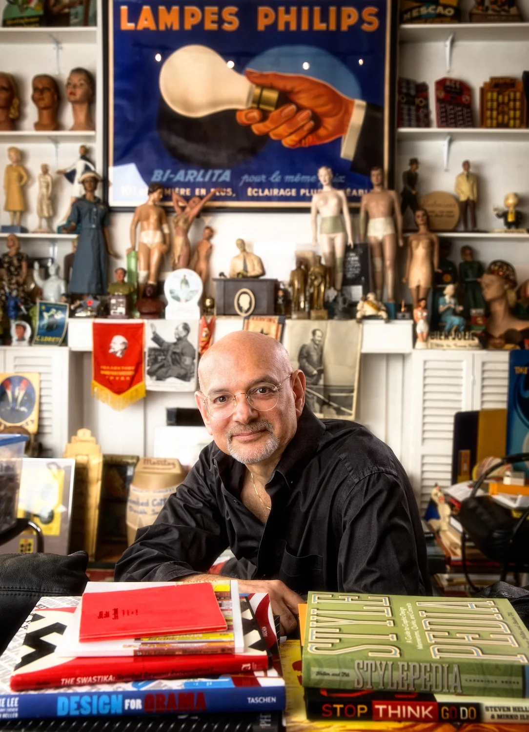 On a recent Saturday morning Steve Heller met me on the steps of his 16th Street studio in Manhattan. I was intent on photographing this icon of design amidst what proved to be the most intense personal collection of objects and books that I had ever seen.
Steve is the latest subject in my graphic designer portrait series. He was the noted art director of the New York Times Book Review for 33 years. He has authored and co-authored over 100 books on design and popular culture. He is a fascinating subject whose charm and accomplishments would be difficult to convey in a single photograph. My biggest challenge was finding a place to put my camera and tripod. I wedged myself into a tiny space between teetering towers of rare books and objects, terrified that I could knock something over and start an avalanche. (Click on photo to enlarge) 