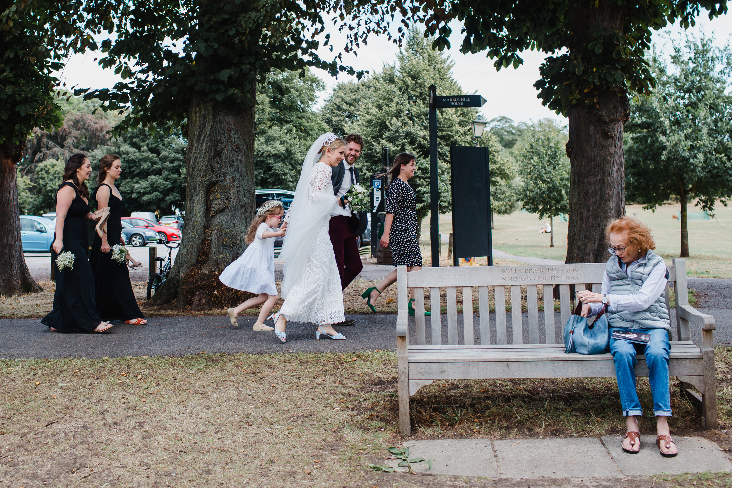a wedding party walks through a park in twickenham while an old lady watches on.