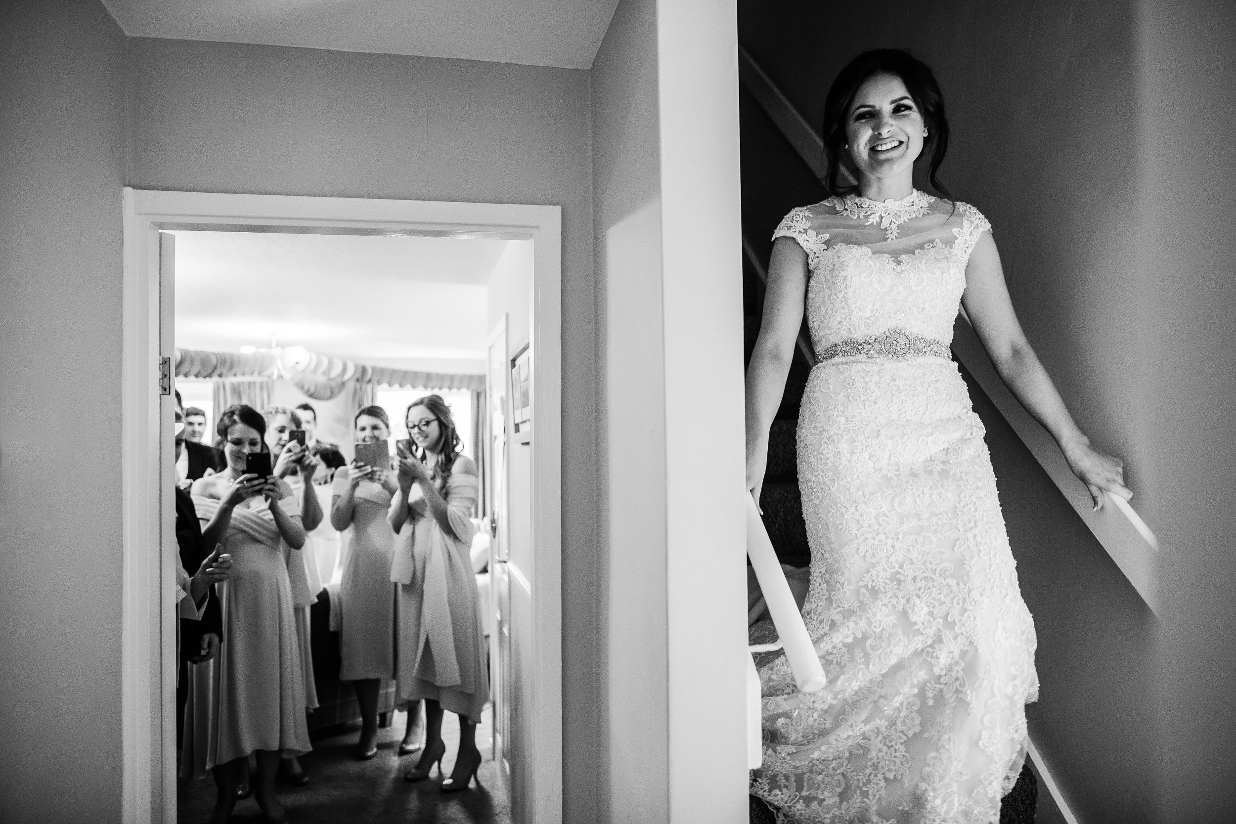 a bride comes down the stairs while her bridesmaids wait for her with phones out