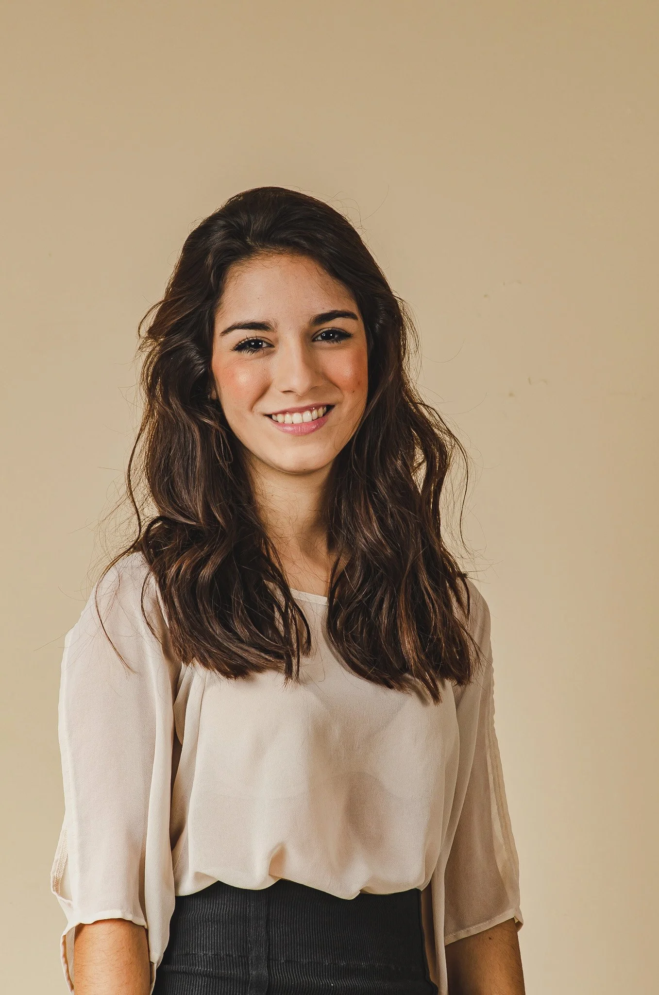 A young woman with shoulder-length brown wavy hair, smiling and wearing a beige blouse and dark pants, standing against a plain light-colored wall.