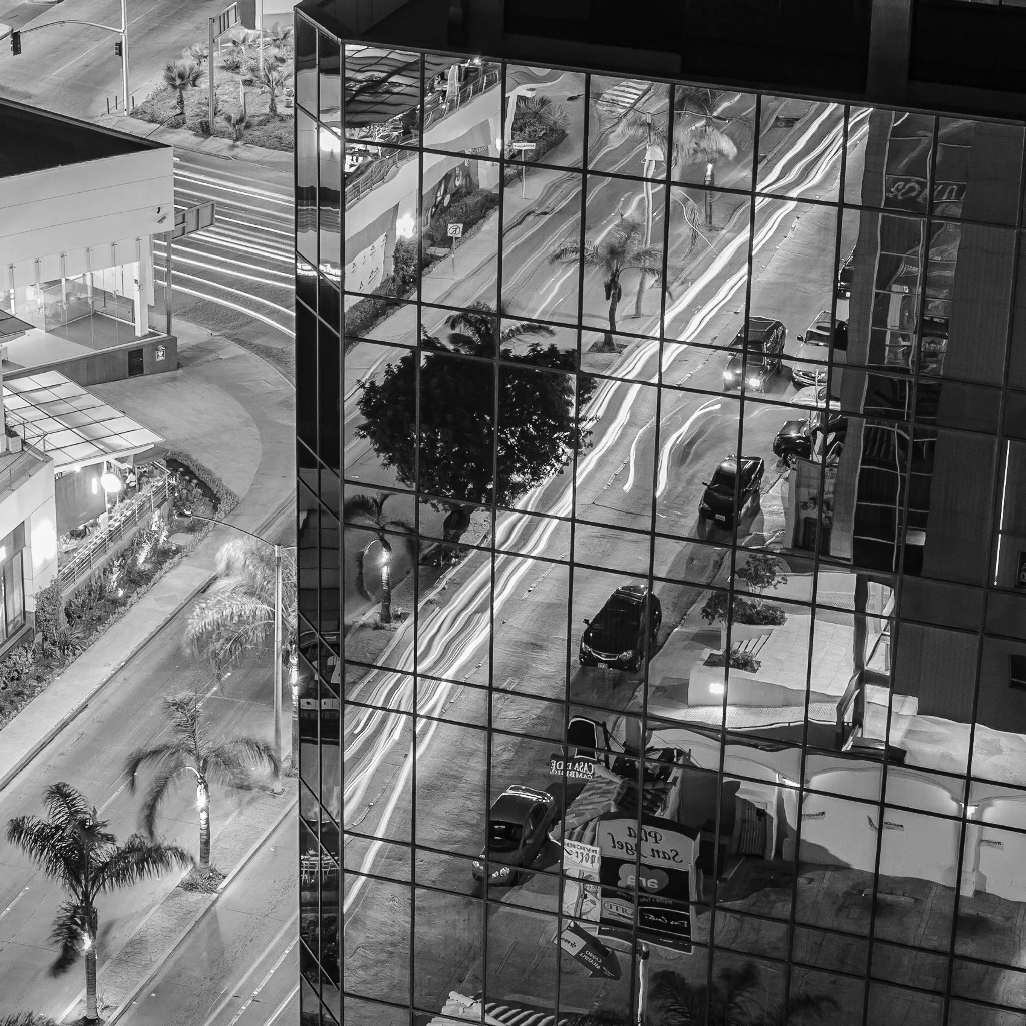 Reflection of urban street scene with cars, trees, and buildings in glass windows of a tall modern building at night.