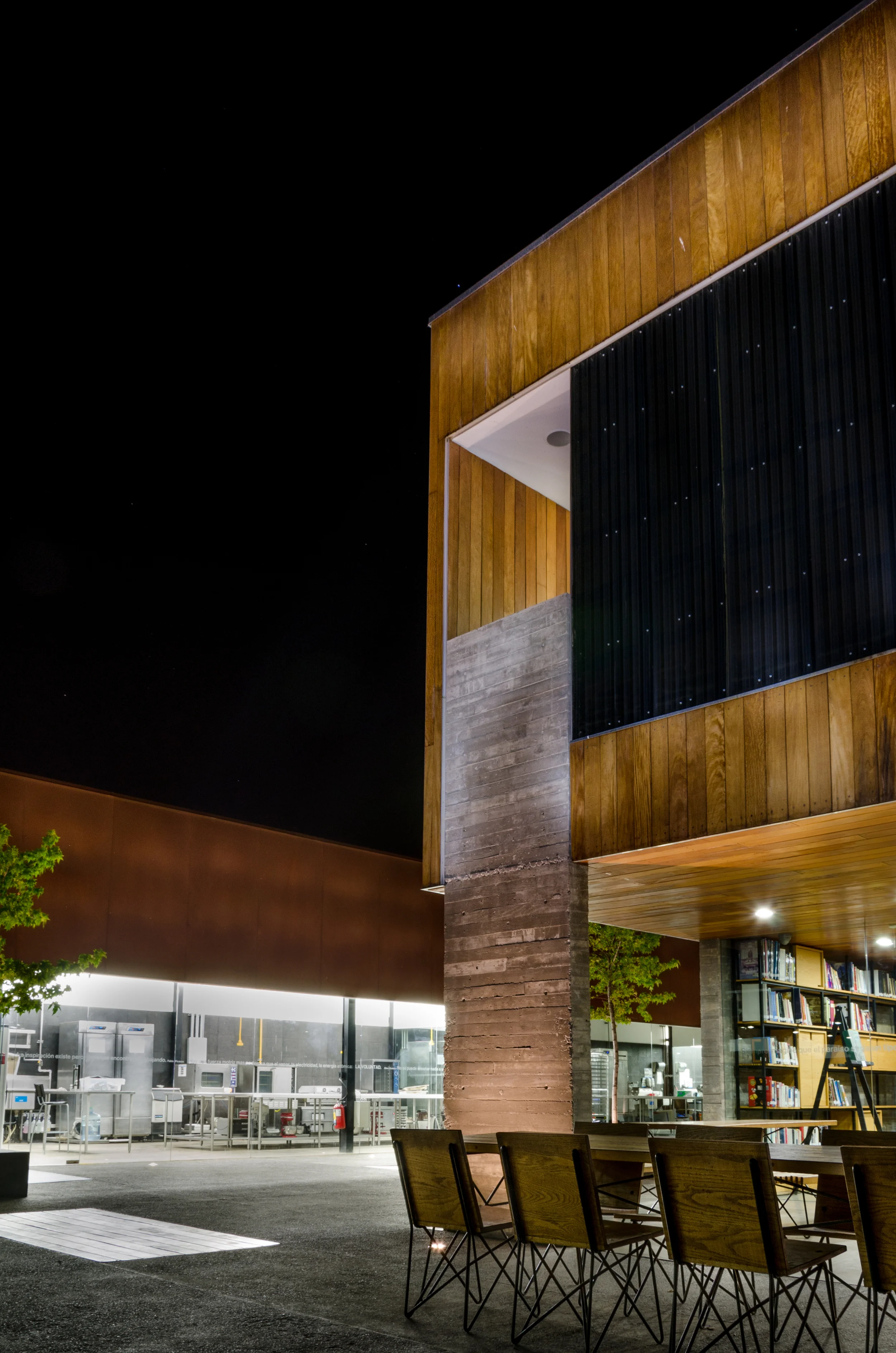 Modern outdoor seating area with wooden and concrete architectural elements, featuring a table with chairs and bookshelves, under a night sky.