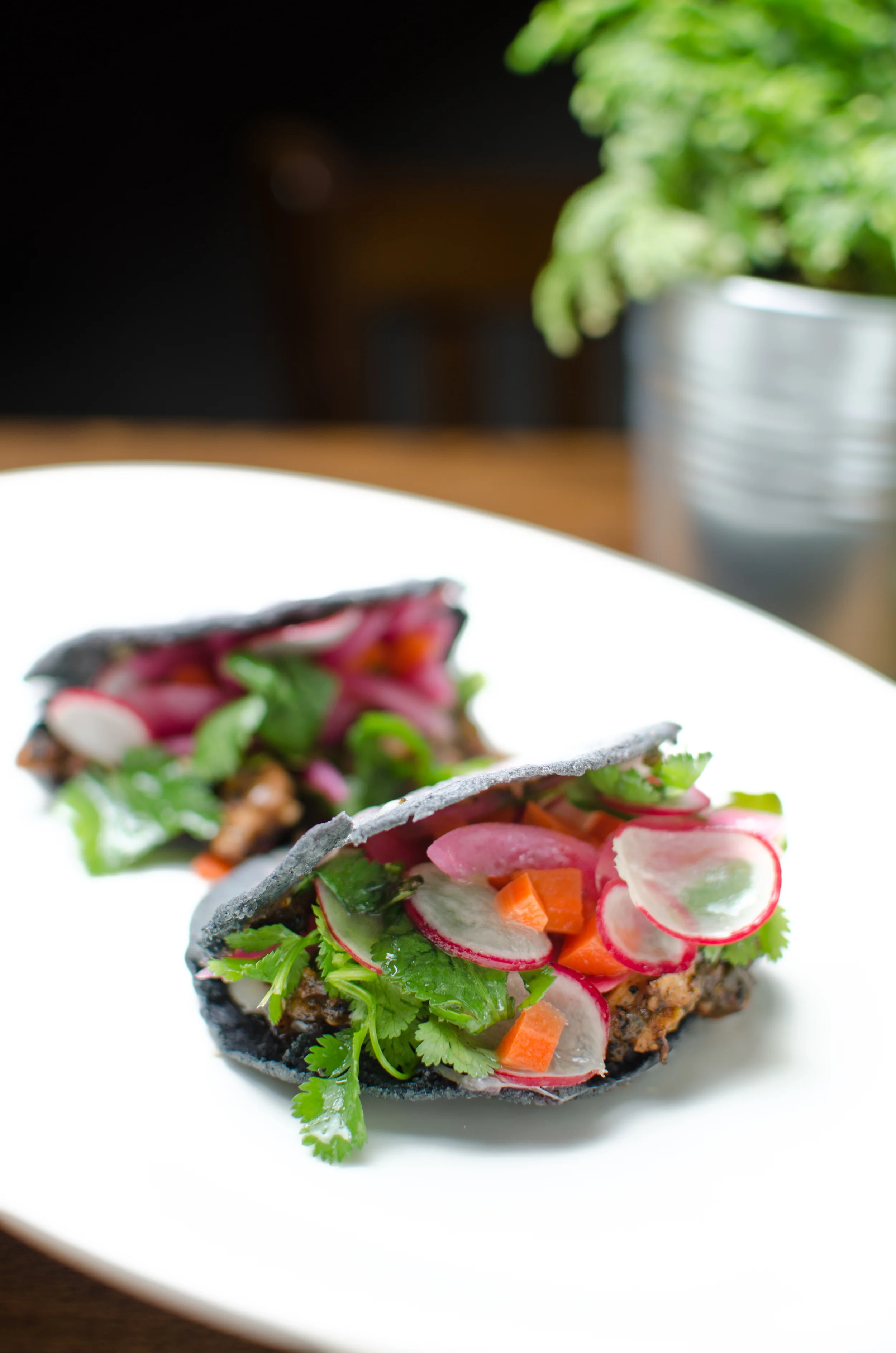 Tacos with blue corn tortillas, topped with sliced radishes, cilantro, and diced orange vegetables on a white plate.