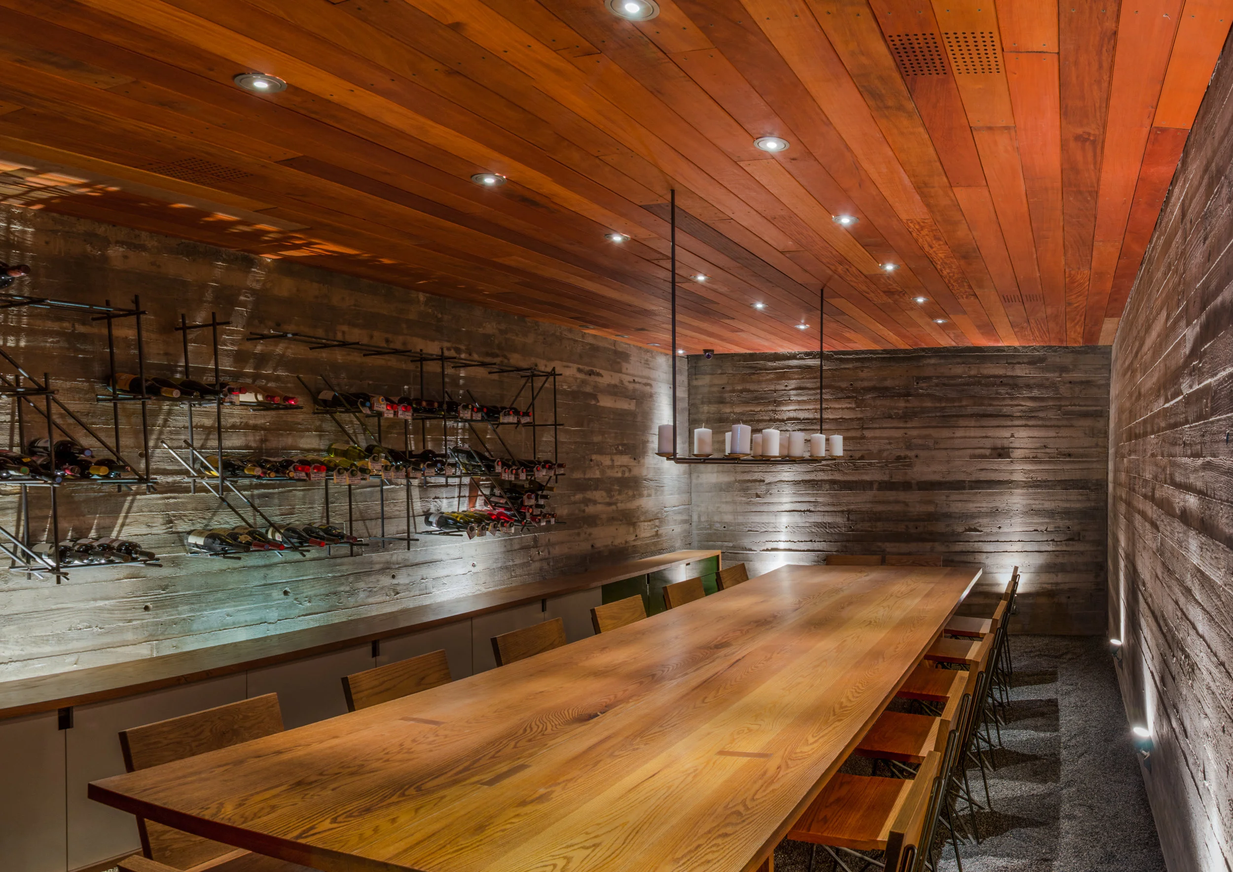 Modern wine cellar with a wooden ceiling and walls, featuring a long wooden dining table, candles, and shelves of wine bottles.