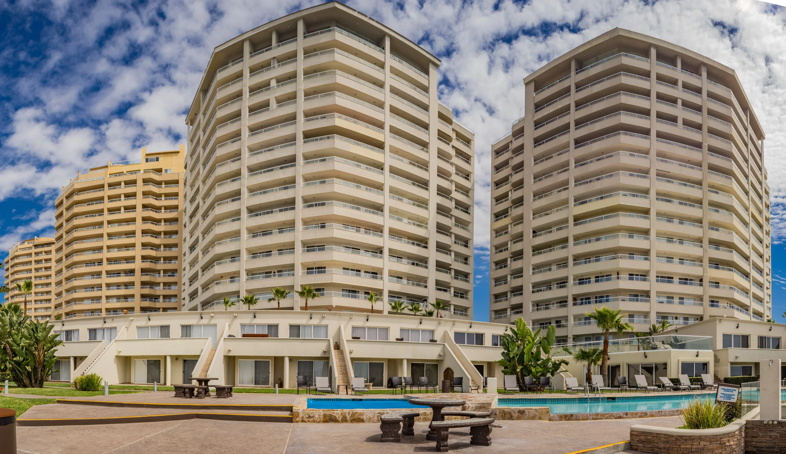 High-rise beachfront condominiums with balconies, outdoor pool, and patio area under a partly cloudy sky.