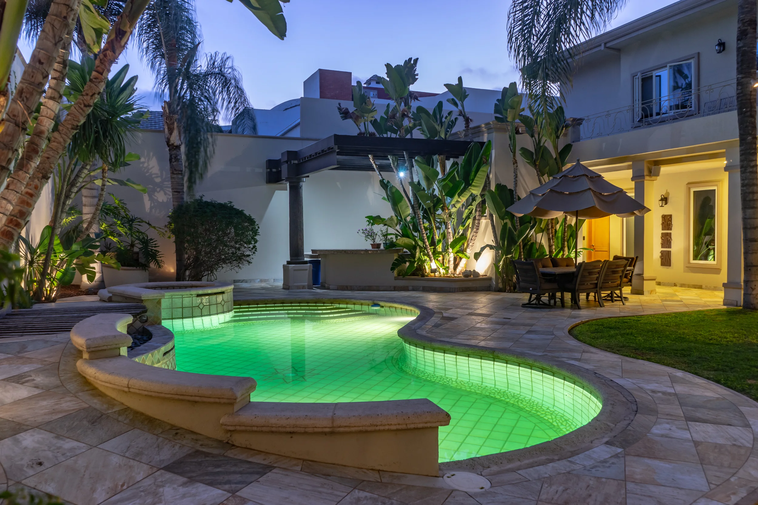 A nighttime view of a luxurious backyard with a glowing green-lit swimming pool, surrounded by tropical plants and palm trees. There is a patio with a dining table under a large umbrella, adjacent to a modern house with large windows and a balcony.