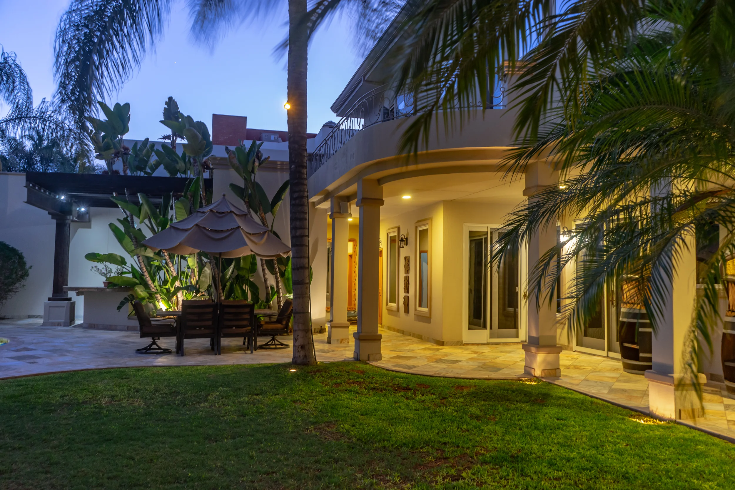 Modern house exterior at dusk with outdoor seating and lush garden, featuring palm trees and ambient lighting.