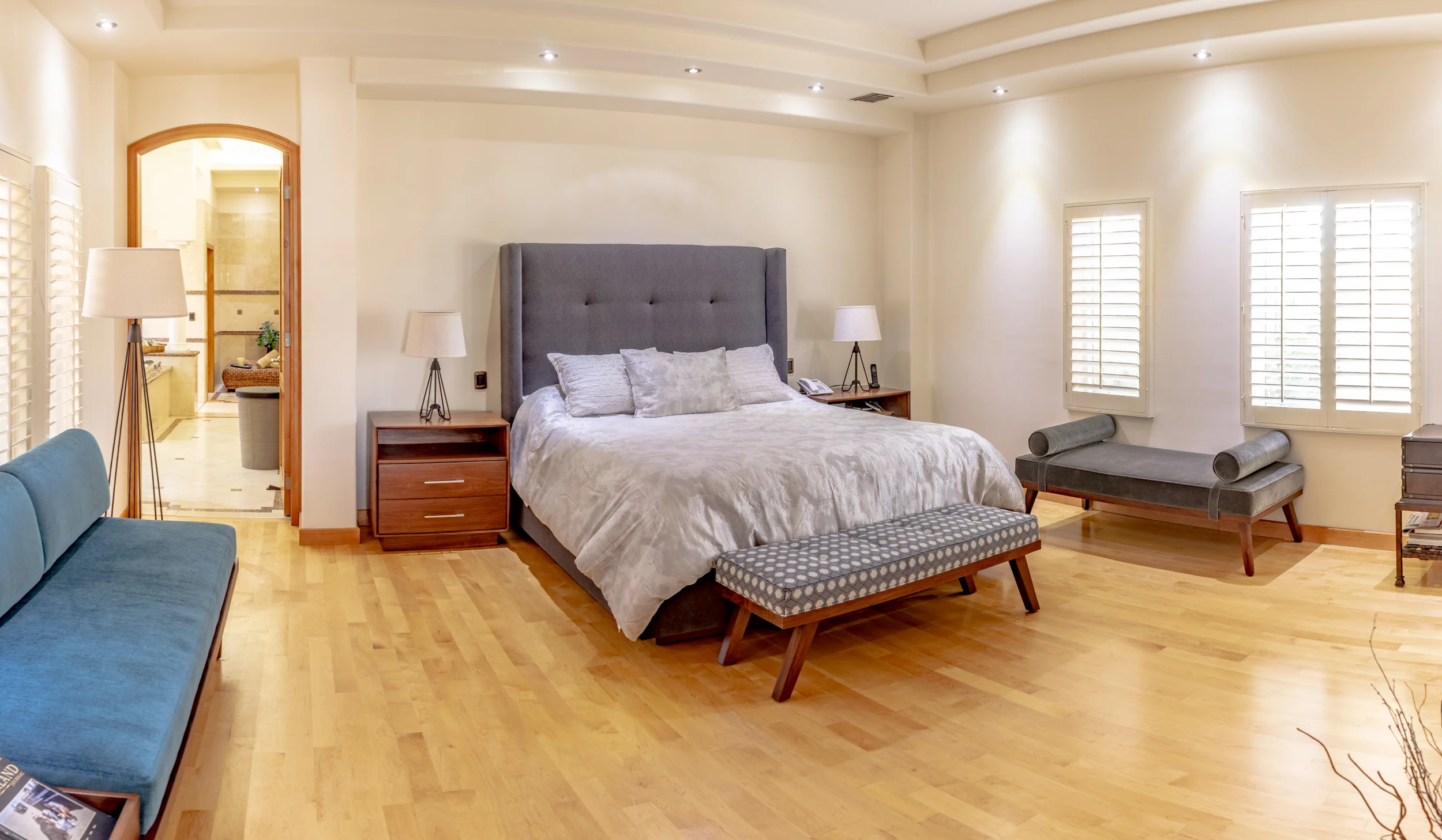 Modern bedroom with a large bed and blue sofa, wood flooring, bedside tables with lamps, and an entrance to a bathroom.