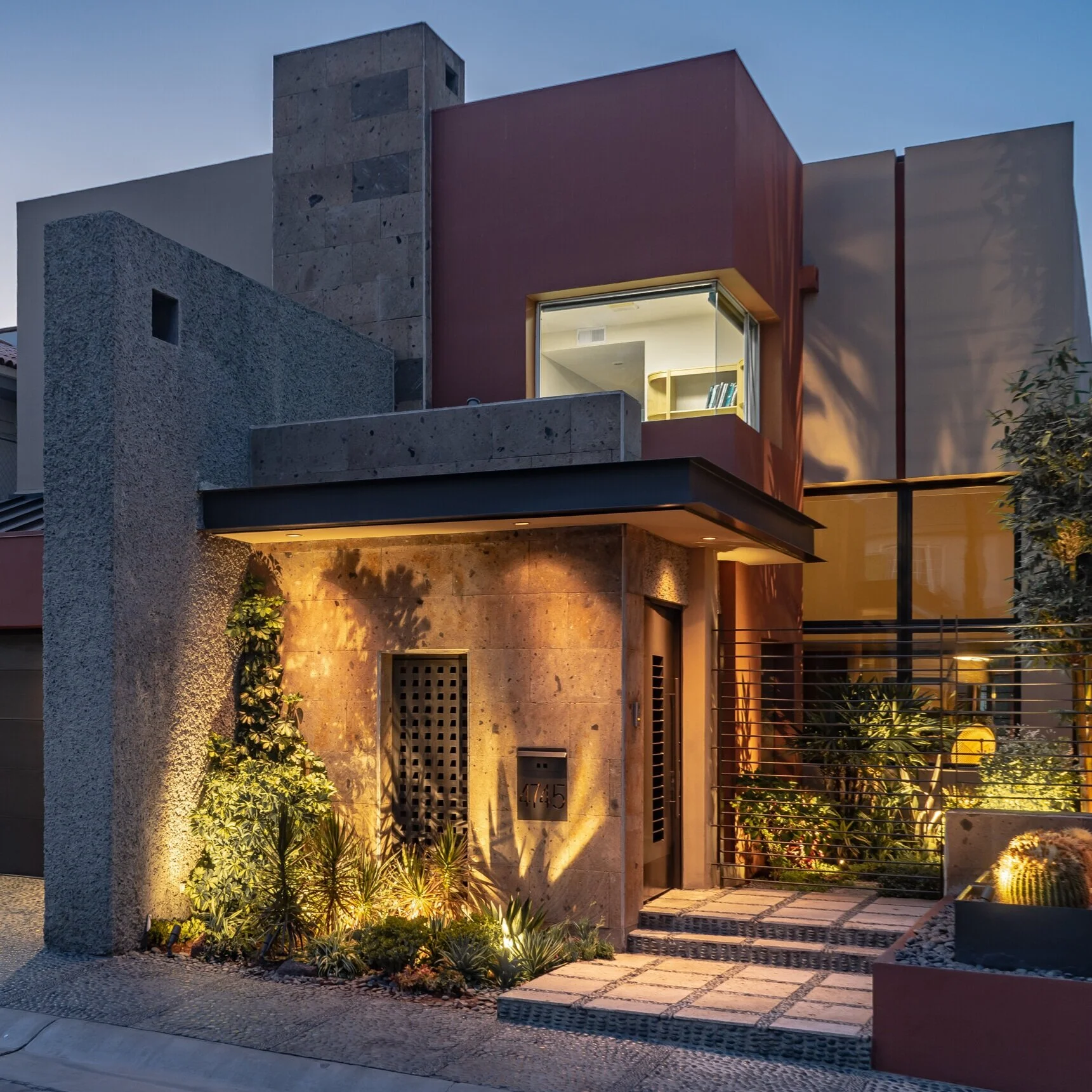 Modern two-story house with concrete and red exterior walls, front entrance with outdoor lighting, plants, and a pathway at dusk.