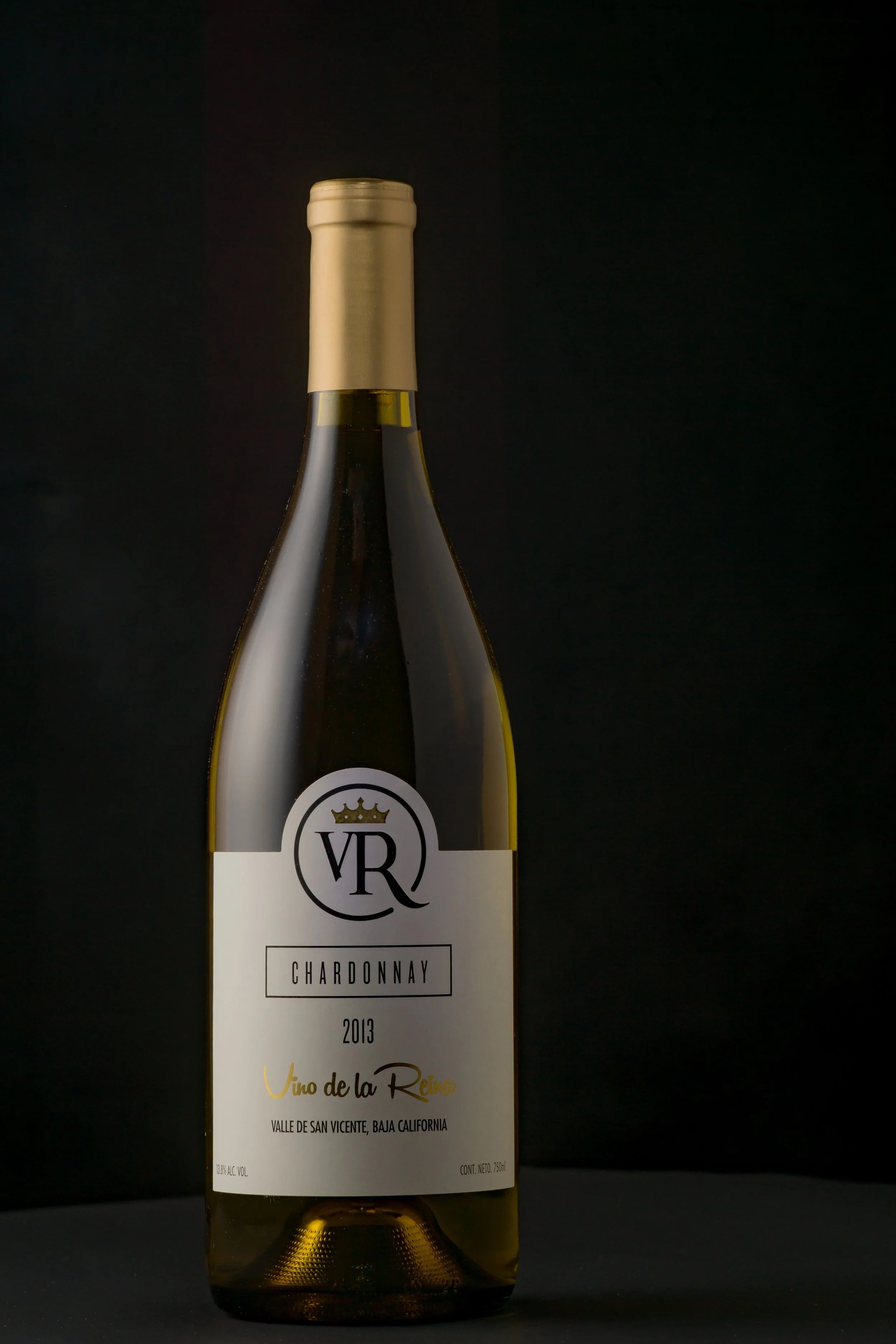 A 2013 bottle of Chardonnay wine from Valle de San Vicente, Baja California, with a gold capsule and a white label featuring a logo with a crown and the initials "VR."